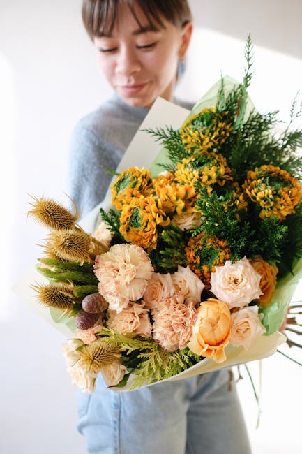 A woman holds a large floral bouquet featuring a mix of fresh flowers including peach and cream roses, bright yellow mimosa, and other colourful blooms, arranged with green foliage. The bouquet is wra