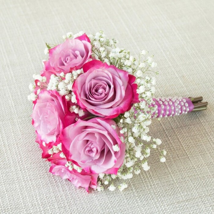 This sophisticated wedding bouquet echoes the serenity and charm of Canonbury's leafy lanes. At its heart, five roses appear almost sculptural, their petals carefully unfurled to reveal a mesmerising gradient from tranquil lavender centres to bold fuchsia fringe-a palette that subtly references the exuberance of East Canonbury's gardens in spring. Feather-light sprays of pure white baby's breath emerge playfully between the rose blooms, offering frothy volume and complementing the rich tones with a whisper-soft contrast. The bouquet is gathered with precision, its stems encased in a smooth, regal purple ribbon, elegantly entwined by luminescent white pearls that spiral downwards, shimmering as they catch the light. Just beneath, the natural green stems peek from their embrace, alluding to the bouquet's freshness and the skilled touch behind every stem. The entire composition sits serenely atop a textured, neutral fabric-perhaps linen or rustic cloth-allowing the vibrant hues to sing out. Viewed from above, the arrangement's rounded silhouette and intricate detailing become clear, making it ideal for a bridesmaid, a flower girl, or a charming centrepiece at a Canonbury gathering. With its blend of modern elegance and timeless romance, this bouquet exemplifies Canonbury Flowers' commitment to exceptional quality and local style.