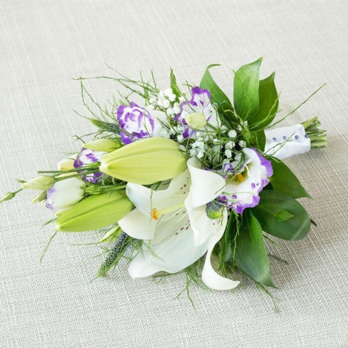 A sophisticated hand-tied bouquet lies diagonally across a light beige, textured cloth, reminiscent of a linen table runner in a Canonbury garden flat prepared for a celebration. The bouquet is composed in an elongated shape, its fullest cluster of blooms resting on the left, gradually narrowing towards the neatly bound stems on the right. Dominating the centre are impressive white Oriental lilies, several of them fully opened so their broad, velvety petals arc outward, framing bold yellow-orange stamens dusted with pollen and the soft scatter of reddish-brown speckles along their inner surfaces. Nestled among these star-shaped flowers are vivid chartreuse-green lily buds, still tightly closed yet clearly swollen with life, adding a sense of anticipation. Woven through the lilies are numerous lisianthus (Eustoma) blossoms, their delicate, frilled petals edged in rich purple that melts into creamy white centres; some are open and cup-like, while others are just beginning to unfurl, suggesting gentle movement and growth. Fine sprigs of white gypsophila (baby's breath) drift between the larger blooms, creating a misty, cloud-like texture. Surrounding everything is a generous collar of greenery: large, glossy dark green leaves give a luxurious, structured backdrop, while slimmer, feathery, and slightly spiky foliage adds lightness and a dynamic edge, like the soft sway of trees along nearby Canonbury Square. The stems are gathered and wrapped in a pristine white satin ribbon, secured with tiny, pearl-like beads that line the handle in a subtle, decorative pattern, unmistakably marking the bouquet as suited to weddings or formal occasions. The lighting is gentle and diffused, with no harsh contrasts, enhancing the fresh whites, soft purples, and vibrant greens, making this arrangement ideal to illustrate refined bridal flowers or special event designs for discerning clients across N1.