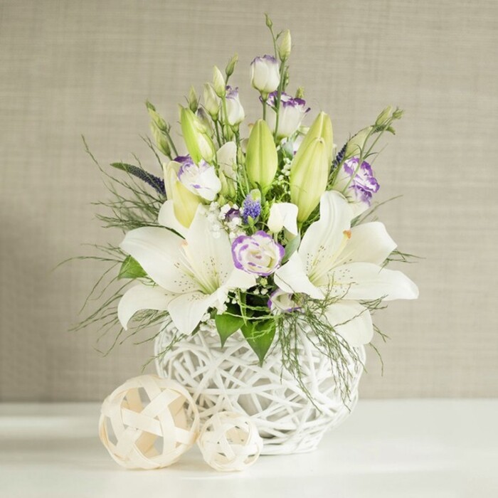 A sophisticated floral centrepiece, ideal for a wedding table in a Canonbury townhouse or restaurant, rests on a smooth white surface, framed by a softly lit beige wall with a faint horizontal linen pattern. The arrangement is anchored by a cluster of pristine white lilies, some trumpet-shaped flowers fully open with visible stamens and a dusting of pollen, others still curved tightly in bud. Their petals, thick yet gently ruffled at the tips, show the slightest shadows of cream along the throat, giving depth and warmth. Around them, stems of lisianthus weave through the design, offering both neat buds and unfurling blooms whose white petals are edged in a whisper of lavender, as though brushed lightly with paint. Here and there, more concentrated deep purple blossoms are tucked between the whites, drawing the eye inward without overpowering the calm palette. Layered amidst these blooms is a mixture of fresh greenery: feathery, fern-like fronds arching toward the sides, slender sprays of foliage adding vertical line, and broader leaves filling the lower portions, all contributing to an airy, garden-inspired structure typical of Canonbury Flowers' style. The bouquet is presented in a distinctive round white vase crafted with an intricate open-lattice design that resembles woven wicker or rattan, giving a modern, sculptural feel while echoing natural textures. Beside it, two decorative woven spheres in pale beige tones rest casually, their curved lines forming a quiet dialogue with the vase's pattern. Natural daylight bathes the scene, highlighting the cool whites, soft purples, and fresh greens, creating a tranquil, contemplative mood suited to weddings, anniversaries, or moments of remembrance across Canonbury.
