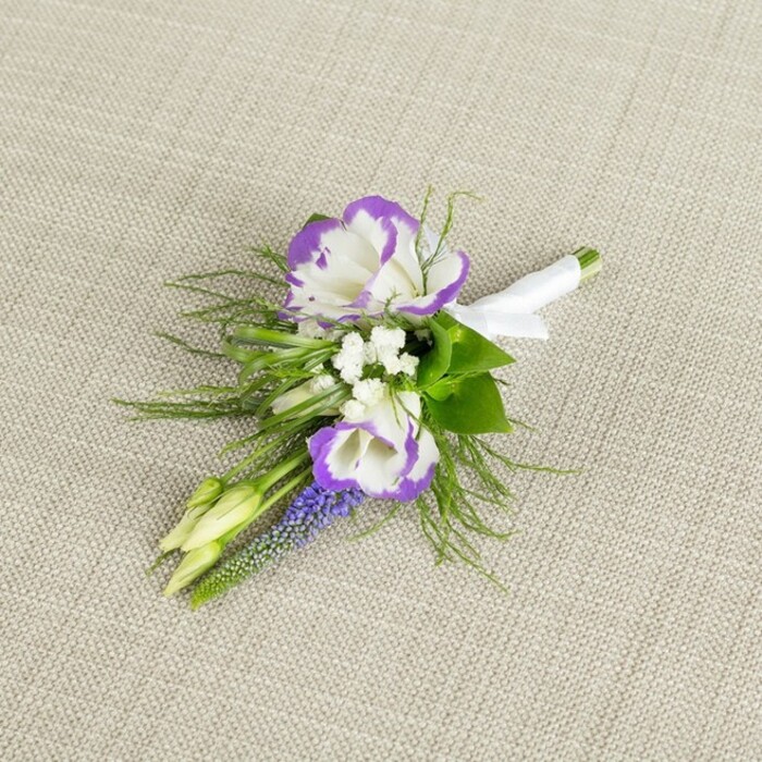 A refined floral boutonniere is photographed in close detail, resting on a softly textured light beige fabric that looks like natural linen, its neutral tone allowing every petal and leaf to stand out clearly. In the centre, two lisianthus flowers unfurl, their creamy white petals tipped with a strong band of deep purple, the ruffled edges creating a sense of movement and gentle drama. Just to one side, a slender spike of muscari rises, composed of tiny, dark purple bell-shaped florets clustered tightly together, offering a more structured, vertical element against the softer curves of the lisianthus. Around these central blooms, small white filler flowers, similar to baby's breath, tuck into the gaps, their fine, star-like petals brightening the arrangement and lending an airy, cloud-like quality. Fine, feathery greenery-evoking asparagus fern-fans delicately around the boutonniere, its light texture softening the edges and adding a freshly picked-from-the-garden feel, like something gathered after a walk through the quieter corners of Canonbury's tree-lined streets. Two pale green buds sit within the design, still closed, giving a sense of freshness and the beginning of something new-perfect for a groom or partner stepping into a wedding day. The stems of all the flowers and foliage are carefully aligned and wrapped in a sleek band of white satin, the ribbon smooth and slightly lustrous, finished with a subtle bow that marks this as a polished, ready-to-wear piece. The lighting is gentle and even, enhancing the soft whites, rich purples, and lush greens, and suggesting an intimate, thoughtful preparation-perhaps arranged earlier that day in the workroom at Canonbury Flowers before being hand-delivered to a nearby townhouse ceremony or a reception space off Upper Street.