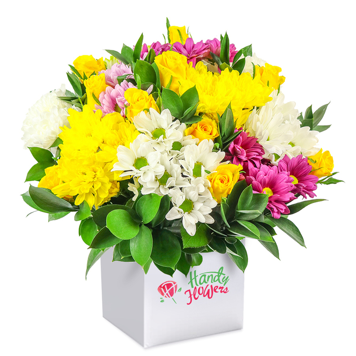 A richly textured, dome-shaped floral arrangement bursts from a sleek white square gift box, carefully photographed against a pure white background so that the colours appear crisp and saturated. The bouquet is densely packed and rounded, giving the impression of abundant garden blooms gathered at their peak and expertly arranged by a local Canonbury florist. Warm yellow tones dominate the piece, especially the large, soft-headed chrysanthemums whose many slender petals create a plush, almost cloud-like surface. Nestled beside them are clusters of yellow spray roses, some fully open to reveal intricate centres, others still in tight buds promising to unfold over the coming days. Punctuating this golden warmth are clean white blooms: small, daisy-style chrysanthemums with bright green hearts and larger, fluffy white mums that introduce lightness and spaciousness within the dense design. Threaded throughout, vivid fuchsia and magenta chrysanthemums add a bold, contemporary energy. Some petals show gentle gradations from deep pink to lighter blush tones at the edges, mirroring the subtle variety one might see in the borders of Canonbury's private gardens. Here and there, petite pink and yellow rosebuds peek out, lending a sense of movement and layered depth. Deep green foliage, likely robust ruscus or similar leaves, surrounds and supports the flowers, its smooth, slightly shining surfaces contrasting beautifully with the soft textures of the petals. The white box vessel is modern and understated, acting as both vase and presentation, making this arrangement suitable for a dining table, office reception, or hallway console. The overall mood is joyful and uplifting, ideal for birthdays, get-well wishes, or a bright thank-you gift delivered to homes and studios around Canonbury, Highbury Fields, or nearby Essex Road.