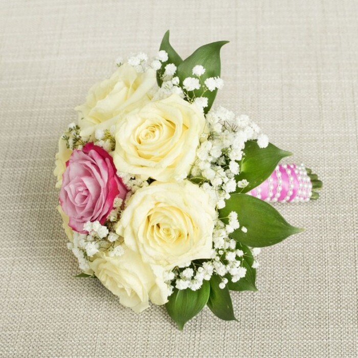 A graceful bouquet, fresh from a Canonbury florist's table, lies nestled on a softly textured cream linen, capturing the refined spirit of Islington's leafy enclaves. Generous, creamy roses-five or six in bloom, their centers tenderly coiled-form the bouquet's luminous core, their subtle fragrance mingling with the gentle aroma of lush greenery. Off-center, a fearless magenta-pink rose claims attention, its luminous petals introducing a modern twist to the traditional palette. Sprigs of baby's breath, fine and cloudlike, are woven throughout, reminiscent of the subtle, fleeting beauty of Canonbury's spring blossom. Surrounding the flowers, a collar of glossy, verdant leaves provides contrast and structure, much as local hedgerows border tranquil garden paths. The bouquet's handle, extending to the right, is bound in a vivid fuchsia-pink satin ribbon, around which iridescent pearls trace a graceful spiral, adding both tactile richness and delicate sparkle. Illuminated by soft, natural daylight, the bouquet evokes feelings of romance and anticipation, making it just the thing for a wedding at Canonbury's elegant venues, a Sutton Place ceremony, or an intimate celebration near Highbury Fields. Each petal, ribbon, and pearl reveals the artistry of Canonbury's floral experts, offering a sensory tribute to love, community, and enduring elegance.