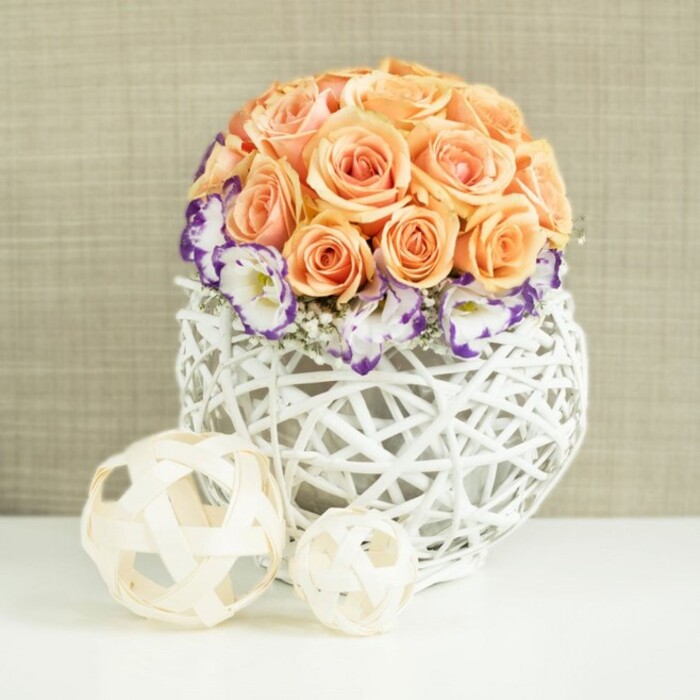 A romantic floral centerpiece is carefully styled on a clean white surface, its understated luxury well suited to an intimate Canonbury wedding breakfast or a stylish townhouse gathering. At the core of the design is a dense, perfectly rounded mound of roses in tender peach, apricot, and soft salmon hues. Each rose appears freshly opened, with layered, velvety petals forming tight rosettes that nestle snugly together, creating an even, dome-like silhouette that feels both lush and orderly. Around the base of this rose canopy, a gentle halo of lisianthus blooms adds intricate detail: white petals finely fringed and dipped in deep violet or purple, as though carefully outlined by hand. Between them, tiny clouds of white gypsophila weave in and out, adding texture and giving the arrangement a light, breathing quality. All of this floral artistry is gathered into a distinctive, bulbous white woven container, its structure crafted from overlapping strands of wicker or rattan, forming a broad, open lattice. The bright, neutral container enhances the soft colours of the blossoms and allows glimpses of stems and shadows to play inside. In front of the arrangement, on the same reflective white plane, two decorative spheres crafted from pale, natural wood strips add a sculptural touch. Their open, intertwined design subtly mirrors the basket's texture, reinforcing the theme of handwoven craftsmanship that you might find in a curated Canonbury interior. A softly textured beige or taupe fabric background frames the scene without distraction. The overall impression is of quiet elegance and enduring romance, an ideal floral choice for wedding tables, engagement dinners, or sophisticated home décor in this leafy North London enclave.