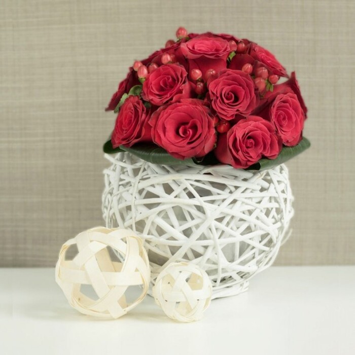 The image shows an elegant wedding table arrangement where deep red roses take centre stage, gathered into a neat, dome-shaped bouquet that almost seems to glow against its pale surroundings. The roses are packed closely together, a tapestry of crimson petals ranging from tightly furled buds to open, velvety blooms that curl softly at the edges. Between them, small sprays of red Hypericum berries are tucked with care, the berries' smooth, glossy surfaces catching tiny highlights and giving the arrangement a jewel-like sparkle. Just at the base of the flowers, a discreet ring of dark green foliage peeps out from underneath, adding a natural outline that enhances the rich reds without distracting from them. This lush floral crown rests in a striking spherical vase made from white-painted wicker or slender branches. The twigs are intertwined to create an airy lattice, leaving gaps that reveal the stems inside and allow light to filter through, giving the piece a contemporary, artisan feel reminiscent of the crafted homeware you might find in independent shops near Canonbury station. The vase is placed on a pristine white tabletop, whose smooth surface reflects just a hint of the shapes above and keeps the composition clean and refined. To the left, two decorative orbs fashioned from pale, natural wood strips sit on the table. The larger, positioned slightly forward, and the smaller, set behind it, repeat the woven texture of the vase but in a warm beige tone, adding a subtle, organic balance. Behind everything, a softly focused wall in light beige or taupe with a gentle texture creates a quiet backdrop. Together, these elements evoke the mood of an intimate Canonbury celebration, perfect for dressing wedding tables, anniversary dinners, or romantic gatherings in local homes and venues.