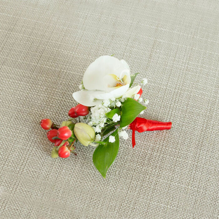 A meticulously crafted boutonniere is photographed from a slightly elevated angle, displayed on a softly textured, light beige fabric that resembles natural linen or burlap, evoking the understated charm of a Canonbury dining table dressed for a special occasion. At its centre, a single white Phalaenopsis orchid bloom opens gracefully, its broad petals smooth and luminous, curving outward like gentle wings. The orchid's core is touched with a subtle wash of creamy yellow and soft orange, drawing the eye to its intricate throat and adding a sense of warmth against the cooler white. Encircling this central bloom is a delicate collar of baby's breath, the tiny white florets forming airy, cloud-like clusters that provide texture and volume without overwhelming the orchid's elegance. Interspersed among the flowers are rich green leaves, some broad and pointed, others slender and elongated, their glossy surfaces hinting at freshness and expert conditioning by a seasoned local florist. Nestled alongside the orchid and baby's breath is a striking cluster of round, polished red berries, reminiscent of hypericum, their vivid colour contrasted by a few softer green buds that introduce a gentle gradient of tone. The stems of the entire arrangement are firmly gathered and wrapped in a bold red satin ribbon, wound smoothly and tied into a compact, tidy knot, with short tails that sit neatly against the fabric. The overall impression is one of poised celebration and refined taste, ideal for a groom, groomsman, or formal guest attending a wedding or winter event in Canonbury, perhaps walking from a townhouse near Canonbury Square to a nearby ceremony, wearing this small yet meaningful piece of floral artistry on the lapel.