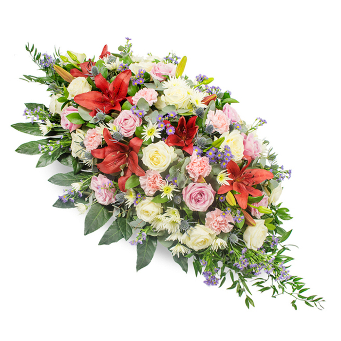 This image shows a refined, double-ended funeral spray laid out against a crisp white background, highlighting the richness of its colours and the complexity of its structure. The arrangement stretches horizontally, widest at the centre and tapering gracefully at both ends, so that the outline feels like a soft leaf or an elegant boat of flowers. Throughout the design, deep red lilies provide striking focal points, their large, star-shaped blooms open wide to reveal speckled petals and golden stamens, suggesting both strength and devotion. These dramatic flowers are interwoven with clusters of roses in gentle hues of cream, white, and pale pink; some roses are tightly furled, while others are fully open, their layered petals forming classic, romantic rosettes. Light pink carnations with frilled edges nestle between the roses and lilies, enhancing the sense of fullness and adding a gentle, almost cloudlike texture. Fine sprays of small white chrysanthemum-type blooms create a bright, clean contrast, while scattered stems of blue-purple thistles bring a modern, slightly wild note, their spiky forms and steely hues standing out from the softer petals. Small touches of lavender or statice-like filler flowers introduce an additional whisper of purple, giving depth without distraction. A lush base of dark green foliage supports and frames the entire piece: broad, glossy leaves suggest salal or laurel, while airy fern-like greens and perhaps a hint of eucalyptus extend beyond the flowers, accentuating the length and movement. The overall effect is dignified and serene, an expertly crafted casket spray of the kind chosen by Canonbury families for funerals at local chapels or crematoria, arranged with the quiet care and experience of a neighbourhood florist.
