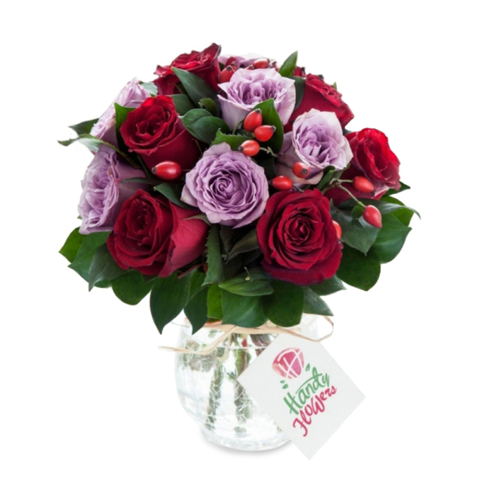 An artfully composed bouquet of red and lavender roses, dense and rounded, photographed on a pure white background to spotlight colour and form. The arrangement balances rich, velvety deep reds - a mix of crimson and burgundy roses - with delicate lilac and dusty purple roses, each bloom fully open with visible, layered petals that suggest freshness and careful timing by the florist. Scattered throughout are small clusters of bright red-orange rose hips, their glossy skins and firm, berry-like shapes introducing a tactile, rustic counterpoint to the softness of the roses. Dark green foliage, likely ruscus or salal, frames the blooms and fills negative space with cool, matte leaves that deepen the overall palette and provide structural support. The flowers are contained in a bulbous, tapering mercury glass vase whose antique silvered and crackled surface reflects highlights and softens the white background; a simple piece of natural raffia or twine is tied around the vase neck for a handcrafted flourish. A rectangular white gift tag hangs forward on the twine, featuring a small stylised rose illustration and an area reserved for a handwritten sentiment. The studio lighting is clean and bright, lifting the true depth of the reds and the gentle mauves while allowing the vase's mottled shine to add a touch of vintage glamour. This composition reads as both romantic and considered - an elegant choice for anniversary blooms, a formal celebration, or a tasteful gift to brighten a home near Canonbury Square or along St. Paul's Road.