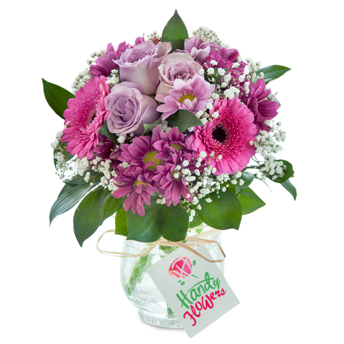 A meticulously crafted bouquet, glowing with the artistic flair of a Canonbury florist, sits serenely against a clean white background-its lush pink, plum, and lavender hues beckoning the eye. At the centre, several elegant, pale lavender roses gently open, their soft fragrance barely hinted at beneath the fullness of the composition. Encircling them, extraordinary hot pink Gerbera daisies command attention with their vibrant colour and dramatic dark eyes, recalling the lively spirit of Canonbury's community markets. Surrounding these focal blooms are clusters of smaller plum and magenta chrysanthemums, each petal arranged to create dimension and movement, much like the skillfully landscaped gardens of Richmond Crescent nearby. Wispy sprigs of white baby's breath intermingle freely, brightening and softening the whole ensemble like the early morning light that streams through Canonbury streets. The greenery forms a verdant base, anchoring the arrangement and providing a fresh, natural contrast. All are presented in a clear glass vase whose distinctive cracked surface refracts light, offering subtle rustic charm. Around the neck, a strand of natural raffia holds a classic white gift tag, open for personalization-a detail that transforms the bouquet into a heartfelt gesture for birthdays, anniversaries, or simply everyday joy, with local delivery across Canonbury and Islington.