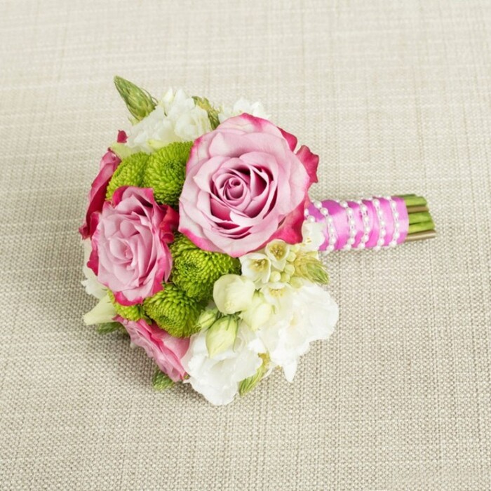On a light tan, textured fabric that looks like finely woven linen laid out in a Canonbury living room, a compact hand-tied bouquet is placed horizontally, as if just set down after a photoshoot or a quiet walk back from a local registry office. The composition centres on three generously ruffled pink roses, with two main blooms in full view and another just tucked behind, each showing a soft transition from pale, almost shell-pink at the inner petals to a deeper, more defined magenta along the outer edges. Their layered petals curl gently outward, creating a plush, romantic focal point. Around and between the roses, small, perfectly domed lime green button chrysanthemums cluster in playful tufts, their dense, tiny florets giving a satisfying, almost cushion-like texture that contrasts with the silken rose petals. Interlaced throughout are sprays of creamy white and soft ivory filler flowers-likely lisianthus or freesia-whose lighter, more fluttering petals add airiness and a sense of movement. Here and there, minute white star-shaped blooms with warm yellow centres suggest waxflower or similar blossoms, adding a delicate, almost confetti-like detail. Subtle snippets of green foliage anchor the design without disturbing its soft, rounded outline. The stems have been cut short and bound tightly, forming a tidy handle. A vivid fuchsia pink satin ribbon is wrapped smoothly around the stem bundle, its sheen catching the gentle, even lighting. Over this polished ribbon, a fine strand of white faux pearls spirals diagonally, like a bracelet clasped around the handle, leaving a glimpse of trimmed green stems showing at the end. The neutral background ensures that the pinks, greens, and whites remain the stars, making this bouquet perfectly suited to a Canonbury wedding, engagement celebration, or baby shower, where elegance and freshness are prized.