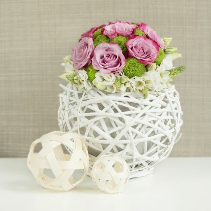 In this serene tabletop scene, a contemporary white woven vase holds a meticulously arranged dome of fresh flowers, evoking the quiet elegance of a Canonbury townhouse dining room prepared for a celebration. The vase itself is spherical and crafted from intertwined white twigs or rattan-style branches, with open gaps that let light pass through, creating a play of delicate shadows on the pristine white surface below. Emerging from this airy vessel, a compact bouquet of roses in soft pink and muted lavender tones takes centre stage, each bloom fully open to reveal intricate swirls of petals edged with slightly deeper pink. Tucked between these romantic roses are bright green button mums, their small, rounded heads adding lively pops of colour and a tactile, almost moss-like texture. Along the outer edges and nestled between the larger flowers, dainty white filler blooms-perhaps small spray roses or similar blossoms-introduce a gentle, frothy softness that breaks up the stronger forms. To the left of the arrangement, resting on the same white tabletop, are two decorative spheres made from light-toned wood or wicker, one larger and one smaller, each with an open lattice structure that mirrors the woven look of the vase. The background is softly blurred and warm beige, reminiscent of linen or burlap, lending a natural, cosy feel without distracting from the bouquet. The lighting is soft and even, highlighting the freshness of the petals and the artful contrasts in texture. Created with the eye of an experienced Canonbury florist, this wedding-style table arrangement feels perfect for intimate receptions near Canonbury Station, relaxed gatherings off Essex Road, or as a sophisticated gift for a local celebration.