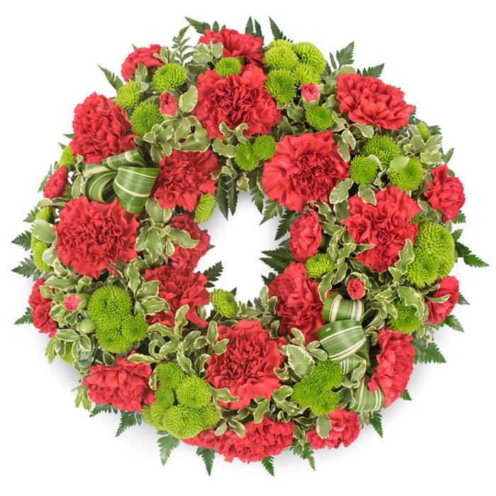 A large, circular sympathy wreath composed entirely of red and green flowers is set against a plain white background, allowing the rich colours and textures to take centre stage. The design forms a perfect, unbroken ring, symbolising the ongoing nature of memory and love, a sentiment often chosen by families in Canonbury for farewell services. Dominating the arrangement are clusters of deep red carnations, each bloom full of intricately ruffled petals that create a plush, velvety appearance. These carnations vary slightly in size and openness, giving the wreath a dynamic, natural quality rather than a flat or uniform look. Interspersed throughout the circle are bright lime green button chrysanthemums, also known as pompon mums, their small, spherical forms packed tightly with tiny florets. Their vivid green colour brings freshness and a sense of light, offering a gentle counterbalance to the solemn depth of the red. Around the flowers and filling the spaces between them is an abundance of foliage. Feathery leatherleaf fern fans out in a rich, dark green, providing a stable framework and a sense of depth, while variegated pittosporum leaves introduce flecks of cream along their edges that catch the light and add visual softness. Woven through the greenery, particularly noticeable along one lower side, is a gracefully looped ribbon in coordinating shades of green, lightly striped, which echoes the natural tones of the leaves while adding an elegant, hand-finished touch. The clean white backdrop and soft shadow beneath the wreath make it appear gently raised, as though ready to be placed at a memorial service, church, or grave in and around Canonbury, offering a respectful and comforting floral tribute.