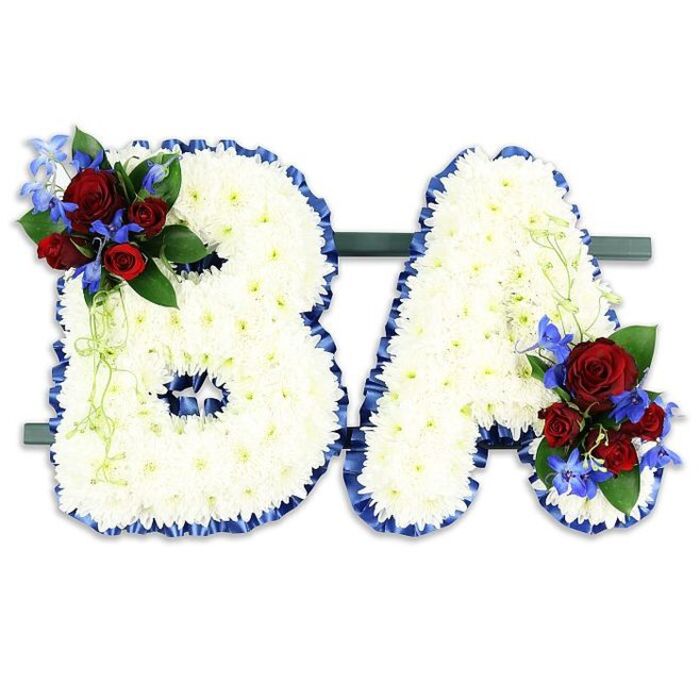 A solemn and meticulously styled memorial piece presents the letters BA side-by-side, each letter formed from a dense mass of pure white chrysanthemum blooms that give the composition a soft, sculptural quality. The chrysanthemums create a continuous, cushion-like surface whose tiny petals catch the light subtly, producing a quiet luminosity across each form. A luxurious royal-blue satin ribbon, pleated slightly along the edges, rims both the 'B' and the 'A', providing a sharp, respectful contrast to the white and reinforcing their shapes with formal clarity. On the upper left arc of the 'B' there is a concentrated floral feature: several richly hued red roses, their petals thick and velvety, set among striking blue delphinium stems whose small blossoms stack like beads of intense blue; together they create a poignant counterpoint of colour and texture. Mirroring this, the 'A' receives a matching cluster at its lower right, where the same red and blue notes are threaded through with dark glossy leaves and pale, descending greenery that softens the letter's line. Both letters are secured on a slender dark-green base that reads as a unified display piece, photographed against a neutral white field so that every hue and contour stands out. For families meeting in Canonbury - perhaps en route from Essex Road or gathering near Canonbury Square - this bespoke lettering is emblematic of a personalised farewell: quiet, carefully composed, and expressive of love, courage, and remembrance.