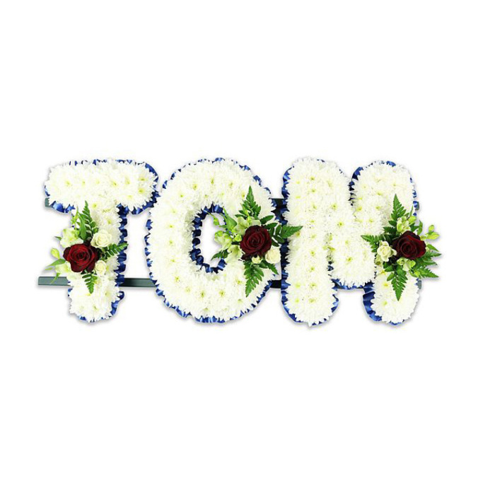 A dignified, personalised floral tribute reads TOM in large, sculptural letters each densely covered with countless small white chrysanthemum-like blooms, their compact petals forming a smooth, pillowy surface that feels soothing to the eye. Every letter's edge is neatly defined by a striking royal blue ribbon, the saturated cobalt line giving formal clarity to the shapes and lending a subtle, respectful flourish. Each of the three letters carries a central decorative spray: a single, velvety deep red rose is placed as the focal point within each cluster, its colour a clear emblem of love and remembrance, while delicate ivory spray roses and small white buds soften the composition and reflect the chrysanthemum tones. Slender fern fronds and lush green foliage are interwoven with the accent sprays, offering feathered texture and a natural contrast that prevents the white surface from looking flat. A dark green or black metal support bar is faintly visible behind the base, indicating the arrangement is designed to stand at a memorial or funeral service. The flowers sit against a bright white background that heightens the colour contrasts and the careful workmanship. As a seasoned local florist at Canonbury Flowers, we craft such tributes with attention to proportion and sentiment, mindful of services held near Canonbury Square, the New River Walk and nearby chapels. The overall impression is one of serene gravity and compassionate care-a handcrafted, sympathetic gesture intended to honour and console those gathered at the farewell.