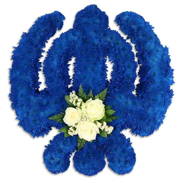 A meticulously crafted Sikh Khanda floral tribute is presented against a smooth, white background, allowing the rich colour and fine detail of the design to stand out clearly. The full Khanda symbol is rendered in flowers: a strong, central double-edged sword, perfectly round chakkar, and two gracefully curved kirpans sweeping outward on each side. Every part is densely covered with royal blue chrysanthemums or similar small-petalled blooms, packed closely so that no gaps show. This creates a thick, velvety surface with a uniform midnight-blue tone, reminiscent of the peaceful evening skies above Canonbury's tree-lined streets. Along the lower portion of the central sword, at the point where the eye naturally rests, a contrasting cluster of white and green blooms adds softness and serenity. Three white roses, their petals spiralling out in elegant layers, sit at the centre of this mini-posy. Around them, fine sprays of tiny white filler flowers, such as baby's breath, bring an airy, cloud-like texture, while arcs of glossy dark green foliage cradle the composition. The interplay of cool blue, pure white, and deep green feels balanced and composed, conveying reverence and a sense of spiritual calm. Gentle, diffuse lighting casts faint shadows that hint at the thickness of the tribute and the careful depth built by the florist. Created as a funeral or memorial piece, this floral Khanda would be fitting for Sikh families in Canonbury marking a farewell, a remembrance ceremony, or a celebration of life. It stands as a respectful symbol of faith, courage, and eternal remembrance, thoughtfully translated into fresh flowers.