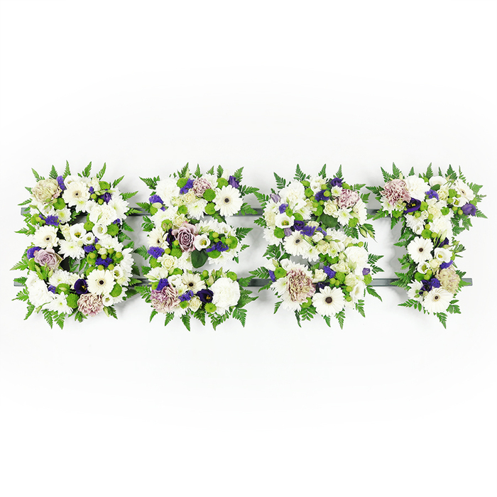 An overhead, horizontal composition spells the word BEST in four densely flowered letters, each letter sculpted from a harmonious mix of whites, lavender, mauve, and fresh greens against a pristine white background. Large, pristine white Gerbera daisies with their darker centres anchor the arrangement, their flat faces offering a calm focal point among layers of ruffled white carnations, soft lisianthus, and compact chrysanthemums that create a variety of petal textures and gentle shadows. Scattered throughout are mauve and lavender roses, their petals curling inward like quiet memories, while deeper purple filler blooms provide discreet accents that lend warmth and depth without overwhelming the serenity of the whites. Feathered fern fronds trace and frame the outer edge of every letter, their airy green fronds providing a soft organic border, and small green button chrysanthemums nestle between blooms, creating pockets of freshness and visual relief. The letters sit on a dark grey structural base that is subtly visible in places, giving the shapes crisp definition and making the word unmistakable from an aerial viewpoint. Under soft daylight or subdued studio lighting the petals show a tender, satin sheen and the overall scent reads as a delicate bouquet of fresh greens and floral sweetness. Designed as a personal sympathy tribute for a memorial or funeral, this arrangement conveys respectful remembrance and handcrafted skill. Prepared in our Canonbury workshop, Canonbury Flowers often crafts similar bespoke letter pieces for families across Canonbury Square, New River Walk and the surrounding Islington streets when words alone are not enough.