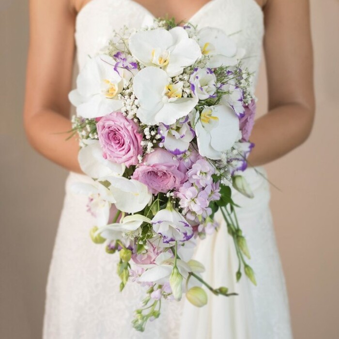 This visually arresting image, set near the characterful terraces of Canonbury, showcases a bride photographed from the chest down, elegantly presenting a lavish cascading bouquet against a softly blurred beige background that hints at neutral stone or historic brickwork. Held within the bride's curved arms, the bouquet unfurls in a graceful shower, led by showstopping white phalaenopsis orchids whose smooth, flat petals gleam with natural light and reveal golden-yellow centers. Nestled among them, large blush-pink garden roses-full and sumptuous-spill their delicate scent, representing enduring romance. Accents of lilac and lavender stock add vertical interest and texture, their slenderness contrasting beautifully with the roundness of the larger blooms. Between these, carefully placed lisianthus blossoms-some edged in a distinctive deep purple-introduce subtle complexity. Clouds of white baby's breath (Gypsophila) and sprigs of fine greenery weave in between, enhancing the bouquet's airy volume and organic descent. The gown's white lace overlays peek from beneath the flowers, reinforcing the composition's sense of sophistication. Altogether, the bouquet's artistic flow and harmonious colour palette reflect a modern yet timeless bridal aesthetic-well-suited to the elegant venues and garden weddings Canonbury is known for. This photograph epitomizes the refined artistry and botanical expertise found at local studios in the heart of Canonbury, offering captivating inspiration for bridal bouquets designed with community and tradition in mind.