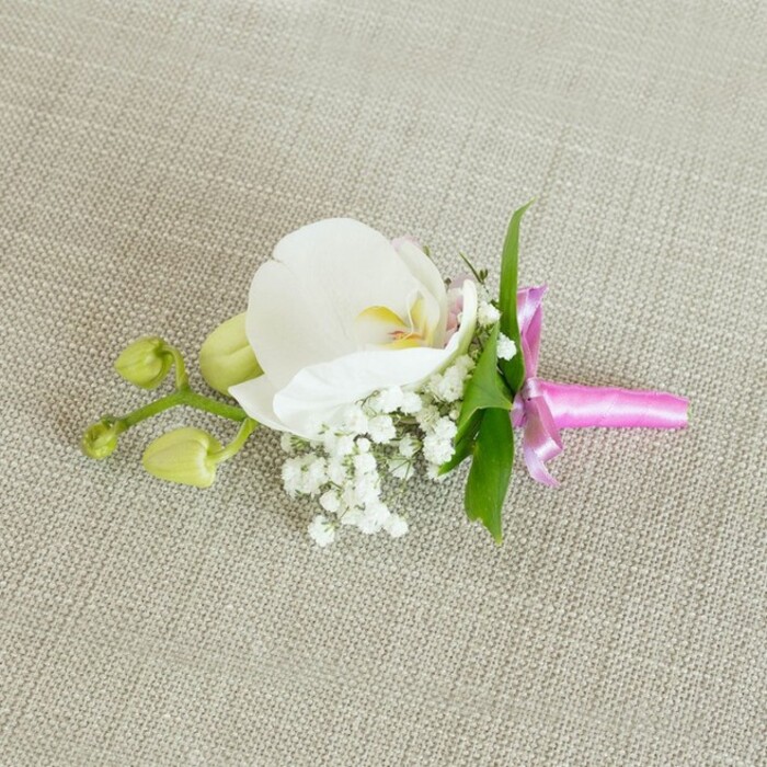 A single white orchid boutonniere is arranged with meticulous care on a light oatmeal-coloured linen surface, the textured fabric acting as a soft, neutral canvas that lets each floral element speak clearly. The Phalaenopsis orchid at the centre is fully open and pristine, its wide petals perfectly symmetrical and slightly cupped as though gently holding the air. Their pure white is softened by the subtle play of light across the velvety surface, while the flower's centre glows with a delicate blend of soft yellow and the faintest blush of pale pink, adding quiet warmth and character. Around the orchid's base, frothy clusters of white baby's breath spread out like a small, airy cloud, the tiny round blossoms and fine stems creating an almost lacy halo. Slender green leaves tuck in behind the flowers, their vibrant, lively hue offering a fresh contrast that reinforces the design's natural elegance. To one side, a fine stem extends out, carrying two tightly furled lime-green orchid buds that lean gently away from the main bloom, suggesting the slow unfurling of new life and the unfolding of a special day - whether it's an intimate Canonbury wedding near St Paul's Road or a smart celebration just off Upper Street. The stems of the boutonniere are brought together cleanly and wrapped in a vivid fuchsia pink satin ribbon. The ribbon is tied into a small, neat bow, its loops and short tails shining softly, adding a pop of modern, celebratory colour that plays beautifully against the white and green. The lighting is soft and even, with no harsh shadows, so the viewer can sense the delicate textures: the orchid's smooth petals, the baby's breath's fine, cloudlike clusters, the crisp grain of the linen, and the satin's gentle sheen. It feels like a timeless piece of floral craftsmanship, the kind a Canonbury florist might prepare moments before it completes a groom's suit, a groomsman's jacket, or a cherished guest's evening attire.