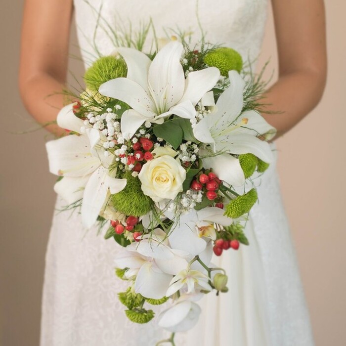 Captured just outside a local Canonbury chapel, this detailed image reveals a bride holding a lavish, cascading bridal bouquet against the ethereal backdrop of an ivory lace gown. Meticulously arranged by an experienced Canonbury florist, several large white lilies stand radiant at the forefront, their elegant trumpet shapes and light freckling offering bridal sophistication. At the center, a creamy white rose swirls tightly, an emblem of enduring love. Throughout the bouquet, bright lime-green cushion mums evoke the spirit of springtime strolls along Canonbury Road, infusing a fresh, contemporary vibrancy. Interspersed, glossy red clusters of hypericum berries add a festive, organic contrast that harks to the rich foliage of Islington's gardens. Wispy baby's breath introduces an airy, cloud-like softness, while sprigs of fine grass and emerald foliage frame the composition, lending a graceful sense of wildness. The bouquet's flowing, teardrop silhouette is elegantly shaped by cascading phalaenopsis orchids, their delicate white petals unfurling downward and lending a sense of movement reminiscent of gentle streams in local parks. The bride's hands and a subtle hint of white satin ribbon anchor the floral display, framed softly by the delicate lace of her dress. The blurred background keeps the focus on the bouquet's striking beauty-a signature arrangement suited for those seeking Canonbury's blend of classic and contemporary in their wedding florals.
