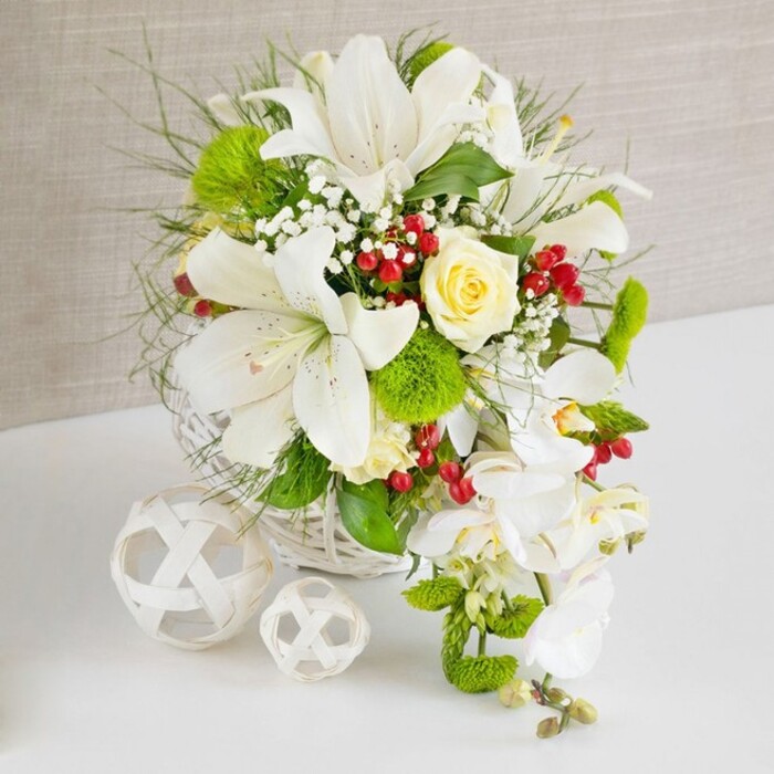 An exquisitely styled bridal-style table arrangement is captured on a clean white surface, the flowers glowing softly against a gently textured beige background that could easily belong to a stylish Canonbury venue. The composition flows from a low, round white basket woven from fine strands that criss-cross neatly, grounding the exuberant cascade of blooms. At the top, large white Oriental lilies open in full, each trumpet-shaped flower displaying smooth, satiny petals with subtle freckles and central stamens dusted in soft yellow, hinting at a light, sweet perfume. Among these lilies, a single ivory-cream rose, its petals tightly layered in an elegant spiral, sits slightly off-centre, lending warmth and a romantic focal point to the design. From the right-hand side of the arrangement, stems of white phalaenopsis orchids descend gracefully, their arching sprays and flat, rounded petals creating a waterfall effect that feels made for a wedding reception in one of Canonbury's historic townhouses. Around and between these signature blooms, a blend of lush greenery provides structure and movement: delicate feathery foliage, slender blades of ornamental grass, and broader, glossy leaves that catch the light. Tucked in for texture are bright green pom-pom flowers, resembling miniature mossy cushions, which contrast beautifully with the smooth orchid petals and polished lily surfaces. Fine clusters of white baby's breath are scattered throughout, creating a misty veil that softens transitions, while clusters of shiny, ruby-red berries add bold accents and a hint of festive charm, perfect for a winter wedding meal near Upper Street. On the left side of the composition, two white decorative spheres rest quietly, woven like airy wicker lanterns, one larger and one smaller, echoing the basket's craftsmanship. The overall effect is one of refined luxury and gentle celebration, a serene yet striking centrepiece for weddings, anniversaries, or intimate gatherings in Canonbury.