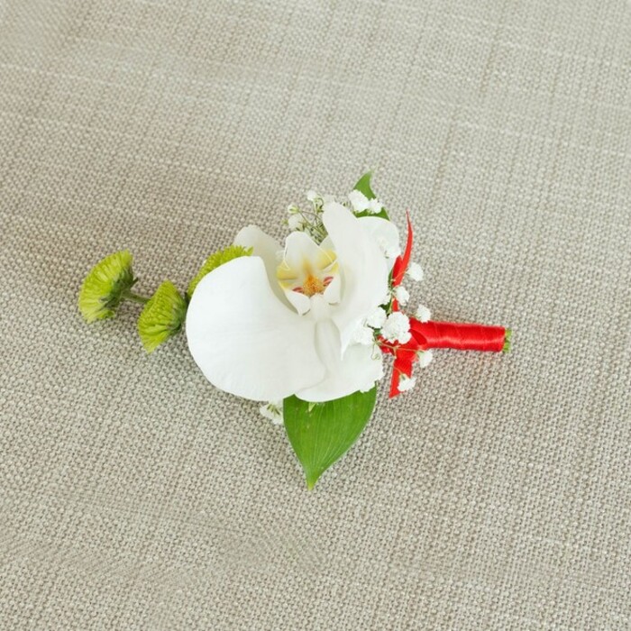 On a softly textured, light beige linen surface that recalls the calm interiors of Canonbury's period homes, a sophisticated boutonniere is carefully laid out, ready for a wedding or black-tie event. At its centre sits a pristine white phalaenopsis orchid, its wide, gently curving petals opening like a serene smile. The orchid's throat is intricately detailed, with soft yellow tones that soften into delicate reddish-orange or light brown markings, giving the flower a subtle, painterly quality when viewed up close. Surrounding the central bloom, fine sprigs of white baby's breath weave in and out, each tiny blossom adding a dusting of texture, like light snowfall resting against the orchid's smooth surface. To the left, two small yet striking lime-green chrysanthemum pom-poms nestle against the composition, their compact, almost spiky petals creating a vivid contrast in both colour and texture, bringing a fresh, contemporary touch. Beneath the flowers, a broad, deep green leaf supports the design like a natural cradle, while a smaller, darker green leaf peeks out near the ribbon, adding layered greenery that feels crisp and alive. The stems are expertly bound in a vibrant crimson red satin ribbon, wrapped neatly and firmly to form a sleek, cylindrical handle. The ribbon finishes in a modest knot or tiny bow, introducing a bold, celebratory splash of colour against the neutral fabric. The even, soft lighting and close framing highlight every detail, from the linen's faint weave to the flower's subtle shadows, suggesting the careful craftsmanship of a local Canonbury florist preparing this boutonniere for a groom walking to his ceremony through the leafy streets near New River Walk.