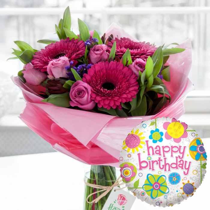 An elegant birthday bouquet and balloon pairing is showcased in gentle natural light, reminiscent of a thoughtful delivery arriving at a Canonbury flat just off Upper Street. The central feature is a tall, clear glass cylindrical vase holding a generous arrangement of fresh flowers, their stems visible through the water and neatly gathered at the base. Dominating the design are large Gerbera daisies in a rich fuchsia pink, their petals flawlessly fanned and framing dark, nearly black centers that add striking depth. Nestled between these bold blooms are clusters of soft blush and mid-pink roses, some in tight bud, others just beginning to open, their layered petals suggesting romantic delicacy and subtle fragrance. Threaded throughout the bouquet are small clusters of deep purple filler flowers, likely statice, which introduce a cool contrast and help give the arrangement a slightly textured, almost meadow-like feel. Lush, bright green foliage provides structure and volume, arching around the flowers and creating a rounded, harmonious silhouette. Around the lower portion of the bouquet, translucent pale pink cellophane or tissue is gathered loosely, forming a soft, crinkled collar that catches the light and emphasizes the feminine colour palette. The vase is finished with a rustic twine bow around its neck, from which hangs a petite, off-white rectangular gift tag adorned with a refined rose emblem, hinting at a premium, carefully chosen present. In the lower right foreground, overlapping the bouquet, a shiny foil Happy Birthday balloon floats into view. Its white background is delicately speckled with grey and scattered with colourful stylised flowers and circular motifs in bright yellow, sky blue, lime green, vivid pink, and royal purple. The words happy birthday appear across the centre in bold pink cursive, mirroring the hue of the Gerberas. The softly blurred indoor setting, likely a bright Canonbury home, keeps the focus entirely on this joyful, ready-to-gift birthday ensemble.