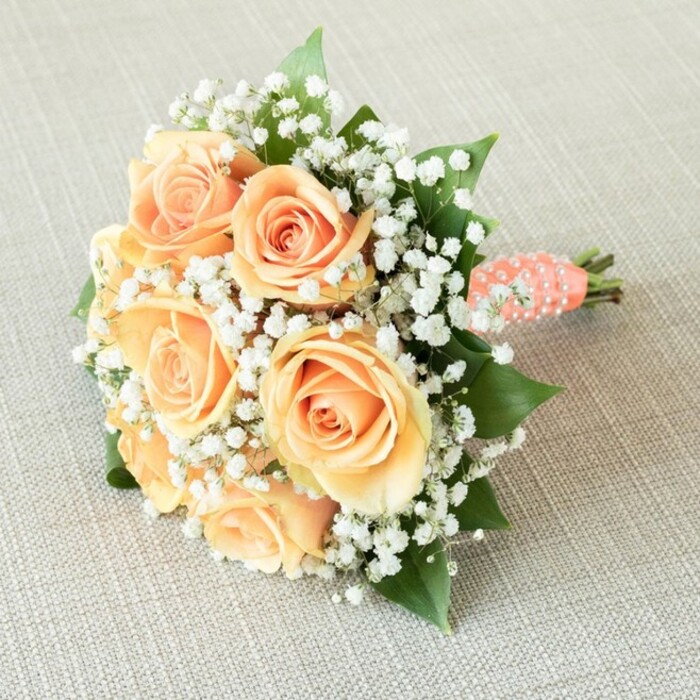 This exquisite bridal bouquet, artfully crafted by a Canonbury florist, captures the tranquil sophistication of London's leafy enclaves. Laid gently on a sandy-beige linen reminiscent of the interiors of Canonbury's timeless townhouses, the bouquet is composed of seven or eight fully-bloomed roses. Their petals transition gracefully from rich peach to pale apricot, each swirl unfolding with tender precision. Wisps of baby's breath, delicate and cloud-like, are scattered among the blossoms, providing a whispered counterpoint of white softness against the bouquet's warm hues. Deep green leaves peek from beneath the roses, evoking memories of the lush foliage lining Canonbury Grove, their color and texture lending depth and grounding to the arrangement. The bouquet is tightly gathered into a rounded, classical pose, signifying careful attention to form and balance. Its stems are bound with a sumptuous coral-peach satin ribbon, embellished by a spiral of lustrous faux pearls that seem almost to glimmer in the gentle north London light. At the base, the cleanly trimmed stems offer a fresh, natural finish. Illuminated by soft, even daylight, the arrangement's subtle color transition, tactile contrasts, and elegantly detailed handle are showcased to full effect. Perfect for local celebrations-weddings held in nearby St. Mary's Church or intimate gatherings along Canonbury Place-this bouquet is as evocative as it is beautiful. It resonates with themes of enduring love and craftsmanship, capturing the essence of a memorable moment curated by Canonbury's own floral experts.