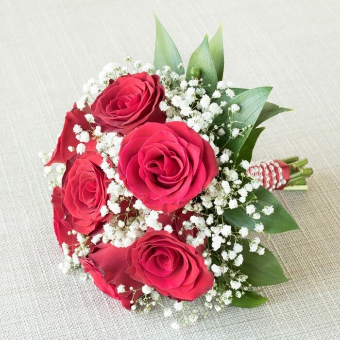 In this elegant Canonbury bouquet, six sumptuous red roses bloom fully, each petal artfully unfurling to reveal a deep, velvety crimson at the heart of the arrangement. The roses, with their classic spiral formation and gentle fragrance, embody passion and devotion-qualities celebrated in North London's vibrant community events and weddings. Interwoven among them are countless dainty white baby's breath flowers (Gypsophila), their delicate clusters echoing the morning mist over Canonbury Square and providing a light, ethereal balance to the richness of the roses. Lush, glossy greenery-possibly ruscus or salal-emerges throughout the bouquet, framing the blooms in verdant freshness typical of the local parks and greenways. The meticulously hand-tied stems are encased in a bold red satin ribbon, elegantly spiraled with a string of reflective pearls, adding just the right note of sophistication and subtle shimmer. The bouquet rests atop a pale, textured surface-likely linen or soft fabric-serving as a gentle, neutral stage that allows the vibrant hues and intricate textures to captivate the eye. Crafted by Canonbury Flowers, this arrangement is designed to convey heartfelt love, romance, and celebration, making it ideal for weddings, anniversaries, or thoughtful gestures to friends and neighbours. The expert composition and skillful craftsmanship shine in every detail, reflecting the artistry and care found in Canonbury's best local flower shops.