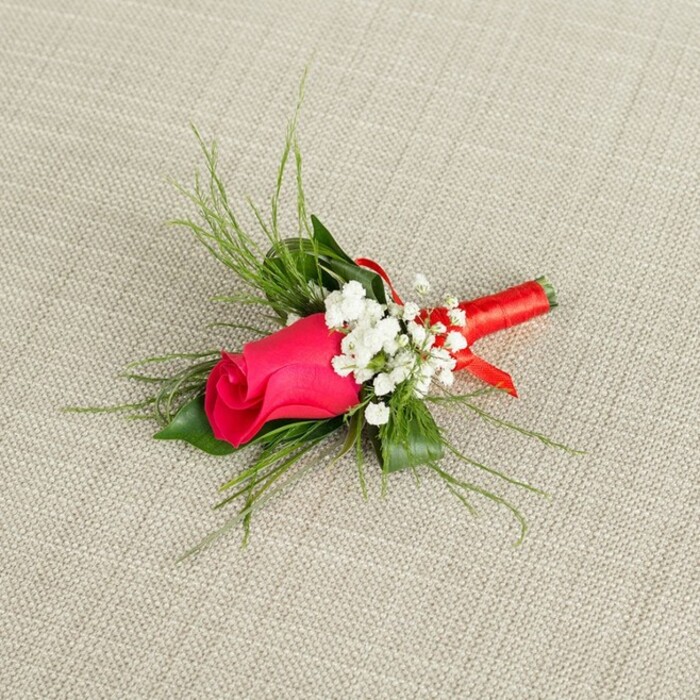 Set on a softly textured, light beige cloth that recalls natural linen or burlap often used in rustic-chic weddings around Canonbury, this boutonnière draws the eye immediately to its central rosebud. The rose is a deep fuchsia or intense crimson-pink, its petals tightly furled and silky, cupping inward to form a compact, elegant shape that feels full of promise, like a moment just before a ceremony begins. Surrounding the rose are airy clusters of white baby's breath, the tiny blossoms gathered in loose tufts that soften the strong colour of the central bloom and lend a romantic, cloud-like effect. Interwoven behind and around these flowers are slender, bright green sprigs of feathery foliage, reminiscent of asparagus fern or fine ruscus, fanning out in arcs that add movement and lightness. A few slightly broader, darker green leaves sit closer to the base, providing a solid, grounding backdrop and making the brighter tones appear even more vivid, as though lit by gentle daylight filtering through Canonbury's leafy streets. The stems of the rose, gypsophila, and greenery are carefully aligned and tightly bound with a vibrant red satin ribbon, wrapped in smooth, overlapping turns to create a polished grip that might end in a subtle knot or small decorative loop. The image is evenly illuminated, highlighting every texture: the sheen of the ribbon, the matte softness of the petals, the finely woven threads of the background fabric. This refined boutonnière, perfect for a groom, best man, or guest at a Canonbury registry office or local church wedding, conveys timeless romance and understated sophistication in a single, thoughtfully arranged stem.