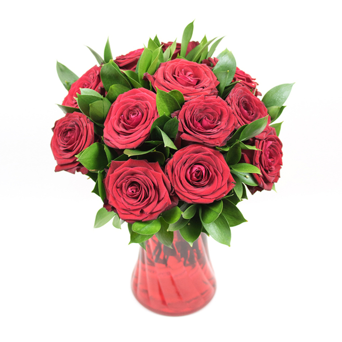 A classic, round bouquet of approximately twelve to fifteen deep red roses stands proudly in a translucent ruby glass vase, set against a bright white background that allows every nuance of colour and texture to show. Each rose is fully open yet still brimming with life, the petals thick and velvety, curling inward in tight spirals at the centre before unfurling into broad, soft edges. The crimson hue is intense and saturated, evoking the warmth of candlelight and the quiet intimacy of shared moments in Canonbury's tucked-away gardens. Thick clusters of dark green foliage thread through the bouquet, most likely ruscus, with its pointed, waxy leaves offering a strong contrast to the softness of the petals. This greenery is not just filler; it builds volume and shape, cradling the roses and giving the arrangement a lush, domed profile that feels both generous and expertly composed. The vase itself is a deep red glass, its body gently curving inwards towards the base before flaring slightly at the mouth. A subtle ribbed or swirling pattern in the glass catches and refracts light, setting off highlights and shadows that echo the layered curves of the blooms above. Because the vase is translucent, the red shifts from darker at the base to brighter near the rim, mirroring the natural variation in the roses' petals. Lit with even, soft illumination, every petal edge, leaf vein, and curve of glass is crisply defined, giving the arrangement a poised, gallery-like presence. It feels perfectly suited to romantic gestures: a surprise anniversary delivery to a Canonbury townhouse, a Valentine's gift waiting on a kitchen table near Newington Green, or a simple yet powerful expression of affection crafted with a florist's practiced hand.