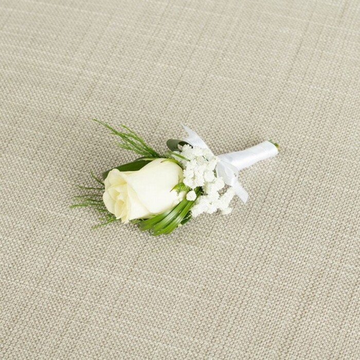 This image presents an elegant white rose boutonniere laid at a gentle angle across a softly textured, light beige fabric background, reminiscent of fine linen or raw silk often used on Canonbury wedding tables. The fabric's subtle diagonal weave creates a discreet crosshatch pattern that sets off the flowers without stealing attention. At the core of the design rests a single creamy white rosebud, its petals smooth, tightly folded, and slightly layered, holding the promise of opening but still in that perfect, pristine moment. A faint hint of green at the base of the rose, where the sepals meet the stem, gives the impression of freshness and recent cutting, as though prepared that morning in a local Canonbury studio. Encircling the rose are airy sprays of white baby's breath, each tiny blossom forming a cloud-like cluster that softens the edges of the arrangement and lends an ethereal, romantic lightness. Complementing these are slender, deep green leaves and a few finer, feathery fronds of foliage, which arc gently around the bloom, framing it and adding a touch of natural movement to the otherwise composed piece. The stems are tightly gathered and wrapped in smooth, bright white satin ribbon, the texture of the fabric sleek and reflective compared with the matte petals and foliage. A small, carefully tied bow sits just below the floral elements, its loops tidy and symmetrical, indicating a piece ready to be pinned onto a groom's jacket, a best man's lapel, or a smart guest's suit for a ceremony near Canonbury Square or along the leafy paths leading down toward Islington. Soft, even lighting bathes the entire boutonniere, accentuating the gentle curves of each petal and leaf, and casting minimal shadow, creating a serene, timeless mood that matches the quiet elegance of a Canonbury wedding morning.
