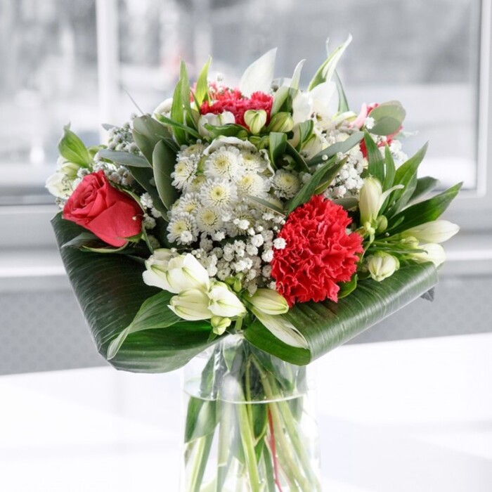 A rich, romantic bouquet of red and white flowers is captured in a moment of calm on a pristine white surface, set against a softly glowing background that feels like a bright Canonbury morning filtering through sheer curtains. The flowers are gathered in a tall, clear cylindrical glass vase, its sides gleaming and transparent, revealing the straight, clean-cut green stems submerged in fresh water. Above the vase, deep red roses with velvety, layered petals open in full rosettes, joined by a large crimson carnation that anchors the front of the display with its densely ruffled, almost frilled texture. Nestled among these passionate reds are creamy white blooms: alstroemeria with slender, slightly flared petals and fine speckling near the center; white daisy-like flowers bringing a soft, open shape; and several white lily buds kept tightly closed, their smooth, elongated forms hinting at fragrance and elegance to come. Tiny clusters of gypsophila, or baby's breath, scatter like delicate snowflakes throughout, making the arrangement feel airy and generous. Encircling the base is a collar of broad, glossy dark green leaves carefully curved and wrapped, while upright stems of eucalyptus-style foliage and pointed green leaves add height, structure, and subtle variations in green tones. The light is even and flattering, picking up the sheen of the foliage and the gentle shadows in the folds of each petal. It is the sort of bouquet a Canonbury Flowers designer might place as a centrepiece for a small gathering off Upper Street, or send out as a same-day delivery to celebrate love, friendship, or a meaningful thank-you, radiating a sense of refined joy and heartfelt intention.