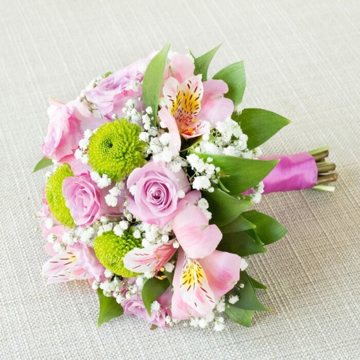 Gracefully displayed on a pale, subtly textured cloth reminiscent of Canonbury's historic homes, this hand-tied bouquet is a symphony of refined floral artistry and gentle vibrancy. Pastel pink roses unfurl at the centre, their spiralled petals exuding timeless romance and delicate charm, seamlessly paired with bright lime green button chrysanthemums whose compact, globed forms bring playful contrast. Airy pink alstroemeria-with slender petals touched by soft yellow and deep speckling-add a light, exotic dimension, echoing the eclectic blend of cultures found around Upper Street. Wispy baby's breath clusters soften and cloud the bouquet, providing a delicate veil that connects each architectural bloom and creates an impression of natural lightness, much like the atmosphere of Canonbury's riverside mornings. The stems, left in their natural green and earthy brown, are gathered tidily and wrapped with an opulent magenta satin ribbon-a sophisticated flourish mirroring Islington's vibrant local palette. Framing the florals, glossy green leaves add depth and lushness, grounding the visual appeal with their freshness. Every element benefits from gentle, diffused lighting, highlighting the bouquet's textural layers and harmonious interplay of colour. Crafted for moments of joy-be it a Canonbury wedding, Park Theatre celebration, or a warm anniversary gesture-this bouquet evokes the promise of springtime, community, and refined happiness. It stands as a testament to Canonbury Flowers' local expertise, transforming seasonal blooms into lasting memories for neighbours and friends alike.