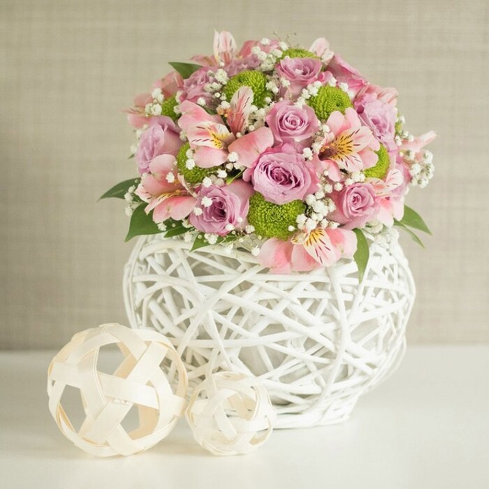 A lush, elegant floral arrangement commands attention in this beautifully lit Canonbury-inspired setting, capturing the essence of refined local design. The bouquet is an abundant celebration of texture and colour, brimming with fully opened pastel pink and lavender roses whose plush, velvety petals promise delicate fragrance and romance. They're joined by trumpet-shaped pink alstroemeria, their petals flecked with unique stripes in yellow and deep rose, adding sophistication and visual intrigue. Bright lime green button chrysanthemums-a playful touch-nestle among the blooms, introducing a lively modern edge. Gossamer sprigs of baby's breath drift between petals, bringing a wispy softness and highlighting the overall lightness of the composition. Interwoven dark green leaves anchor the bouquet, adding natural depth and texture. This vibrant ensemble is gathered in a striking white vase, its open, basket-like latticework reminiscent of bespoke homewares found along Canonbury's local shops. The vase's rounded and substantial form supports the arrangement with confidence, poised on a spotless white surface that sets off the display magnificently. Beside the vase, two ornamental spheres-crafted from pale rattan or bamboo-are strategically placed, their light, woven design echoing the texture of the main vase and adding a minimalist, contemporary flair. Behind, a soft, textured beige background gives the scene a serene, welcoming ambiance without distraction. Altogether, the image evokes the spirit of sophisticated wedding tablescapes and special occasion settings in Canonbury-perfect for anyone seeking beautiful, fresh arrangements that combine classic blooms with artistic modern decor.