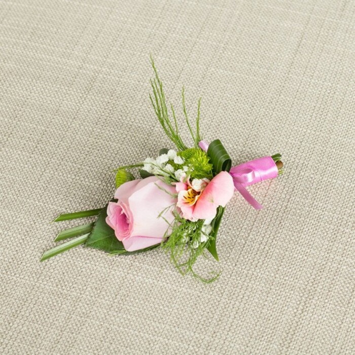 Diagonally placed on an off-white, woven linen surface that echoes the tranquil tones of Canonbury's leafy lanes, this boutonniere is a miniature ode to skilled floristry and gentle romance. At its luxurious core rests a single, perfectly formed pink rose, the petals just beginning to unfurl-a sign of freshness and the anticipation felt on the morning of a wedding. Surrounding the central bloom are airy sprays of white baby's breath and tufts of green cushion mums that recall the lush quiet of local gardens. An additional peach-toned alstroemeria graces the arrangement, its ruffled petals dipped in soft yellow and decorated with subtle freckles, lending further nuance and warmth. The foliage alternates between slender, upright sprigs-perhaps asparagus fern-and broader, elegantly folded leaves in a deep, glossy green, their arrangement forming a natural yet meticulously structured base. A wide, pliant leaf is tightly wrapped around the gathered stems, all secured by a luminous fuchsia satin ribbon tied in a small celebratory bow. Gentle daylight illuminates the intricate folds and subtle colour gradations, casting delicate shadows on the nubby texture below. This floral accessory, ideal for a Canonbury wedding or special celebration, encapsulates both artistry and authenticity. Whether destined for a groom's lapel or a guest of honour, its harmonious composition and thoughtful detail speak to Canonbury Flowers' commitment to local craftsmanship and timeless sophistication.