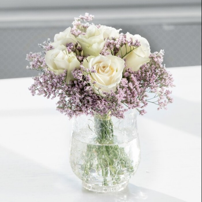 A carefully composed bouquet of creamy white roses and delicate lavender statice sits poised in the centre of the frame, the work of a florist who understands both structure and softness. The roses, about a dozen in number, are at that perfect stage of opening where each bloom is full yet still slightly cupped, their velvety petals overlapping in graceful spirals. Their colour is a gentle cream with an ivory cast, catching the light so that the outer petals seem almost translucent at the edges. Threaded in and around these larger blooms are generous sprays of lilac-tinted statice, each stem branching into clusters of tiny, papery flowers. These small blossoms create a soft haze of lavender purple, diffusing the strong forms of the roses and adding a gossamer texture, like morning mist along the quiet streets of Canonbury Square. The arrangement is gathered in a tall, clear cylindrical glass vase, simple and modern in shape. Through the glass, the viewer can see the green rose stems, some straight, some slightly curved, all cleanly trimmed and immersed in fresh, clear water. The water line and the subtle refraction of the stems lend a sense of coolness, as though the bouquet has just been placed there after conditioning in a florist's sink. The vase stands on a pristine white tabletop, which reflects soft hints of green and cream beneath the flowers. In the background, a softly blurred interior suggests a bright, airy room, lit by daylight filtering through a large window, perhaps in a townhouse just off Upper Street. The gentle illumination highlights every petal edge and statice floret without harshness, creating a tranquil, elegant mood suited to a housewarming gift, a peaceful sympathy tribute, or a calm focal point in a contemporary North London living space.