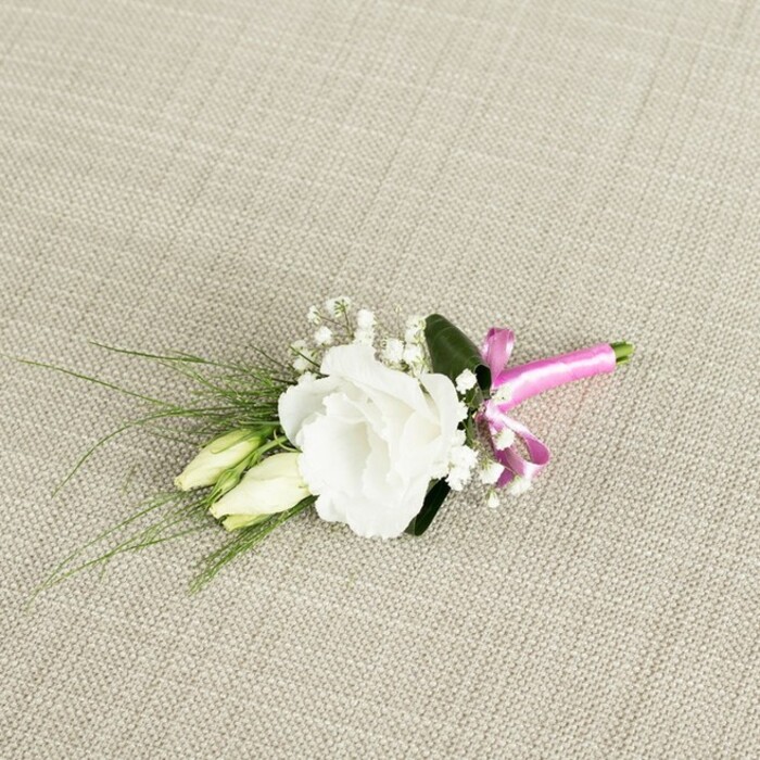 A delicately composed boutonniere is photographed from a slightly elevated angle, resting diagonally on a pale beige, linen-like fabric whose fine weave and subtle texture recall the soft furnishings of a Canonbury townhouse drawing room. At the centre of the arrangement is a fully bloomed white lisianthus flower, its many ruffled petals layered in intricate folds that create a rounded, cloud-like shape. The petals move from a purer white at the edges to a faintly creamier tone near the heart, where shadows pool softly in the interior. Just to one side of the main bloom, two lisianthus buds, still closed and elongated, nestle close; their creamy, pale green outer petals are smooth and taper gently to rounded tips, suggesting potential and new beginnings. Threaded around and between these main elements are delicately branching sprays of gypsophila, each tiny white flower poised on thin stems like clusters of miniature stars, adding a light, airy veil to the design. Emerging from behind the central flower are several slender, grass-like blades of deep green foliage that fan out gracefully towards the lower left, creating a gentle sense of movement and balance. A broader, darker green leaf sits quietly beneath the lisianthus bloom, giving depth and a structured backdrop to the softer forms. At the base, the neatly trimmed stems are bound with a vivid fuchsia satin ribbon, tied into a precise, jaunty bow whose lustrous surface catches the light and introduces a modern, celebratory splash of colour. The overall impression, captured in soft, even lighting, is one of understated elegance and fresh simplicity, an ideal floral accent for a groom or guest at a Canonbury ceremony, perhaps walking from New River Walk to a nearby registry office or intimate reception.