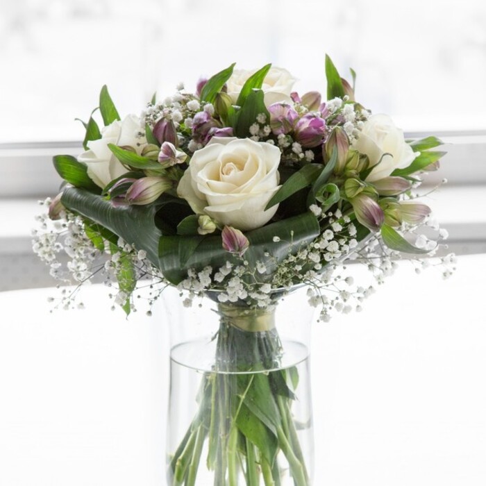 A sophisticated floral display of white and lavender blooms is captured in soft, diffused daylight, as though resting on a table in a bright Canonbury kitchen with light streaming in from a nearby sash window. The arrangement is cradled in a simple, clear cylindrical glass vase, where the long green stems are visible, trimmed with precision and splaying slightly in the water to support the generous, rounded dome of flowers above. At the centre, creamy white roses are in full, luxuriant bloom, their petals unfolding one over another in smooth, velvety layers that suggest both tenderness and timeless elegance. Among these, clusters of alstroemeria stretch upwards and outwards, their slender buds and half-open flowers revealing petals shaded in soft lavender and muted purple, with fine streaks of deeper colour that run towards the throat of each bloom. Interlaced between the more substantial flowers, sprigs of baby's breath float like tiny white clouds, scattering a constellation of miniature blossoms that lend lightness and a gentle, ethereal quality. Surrounding everything, lush green foliage offers a strong yet graceful framework, with broader, darker leaves juxtaposed against more delicate stems, enhancing the natural texture and giving the arrangement a sense of movement. The bouquet feels calm and contemplative, capturing that tranquil moment just before guests arrive for a small gathering off Upper Street or when a thoughtful delivery is placed on a hallway console in a Canonbury flat. Subtle hints of a sweet, garden-fresh fragrance seem to rise from the scene, echoing spring mornings in nearby Highbury Fields. Crafted with the practiced eye of a skilled local florist, the composition works beautifully for expressing admiration, offering comfort during a difficult time, or creating a peaceful focal point in a cherished home setting.