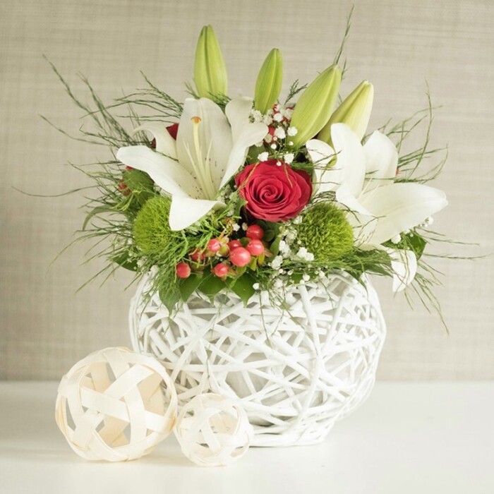 The image presents a beautifully composed floral centerpiece that would sit perfectly on a wedding table in a Canonbury townhouse or a chic restaurant near Upper Street. At its heart is a round, snow-white vase with a distinctive open-weave design, the crisscrossed bands creating a hollow, basket-like form that allows light to play gently through its gaps. This sculptural container cradles an abundant display of fresh flowers and foliage. Majestic white lilies rise proudly from the centre, their satin-like petals opening to reveal soft speckles and long, golden stamens that catch the light. Some lilies remain in bud, tall and tapered in a cool green tone, indicating the arrangement's freshness and the promise of continued flowering long after it is placed on the table. Among the lilies sit deep red roses, including a central bloom whose tightly furled petals swirl inward like a perfect spiral, introducing a romantic focal point ideal for wedding celebrations. Nestled at different heights are bright green spherical blooms, likely green dianthus or chrysanthemum pom-poms, whose dense, fine petals offer a playful yet refined texture, echoing moss or soft velvet. Scattered delicately throughout the bouquet are clusters of tiny white baby's breath, lending an airy, almost cloud-like softness around the stronger shapes. Small bunches of red berries emerge between the petals and leaves, suggesting hypericum and hinting at seasonal festivities without fixing the design to a specific holiday. The greenery is varied and generous, with slender, wispy fronds and deeper, broader leaves creating layers of depth and a rich forest backdrop that makes the white and red flowers glow. The vase rests on a smooth white surface, with two smaller decorative woven spheres in pale natural tones set slightly to the left, subtly balancing the scene and echoing the vase's texture. Behind, a muted beige wall with gentle texture keeps the focus on the arrangement itself. The overall atmosphere is one of understated luxury and intimate celebration, perfectly suited to Canonbury weddings, anniversaries, or elegant dinner gatherings.