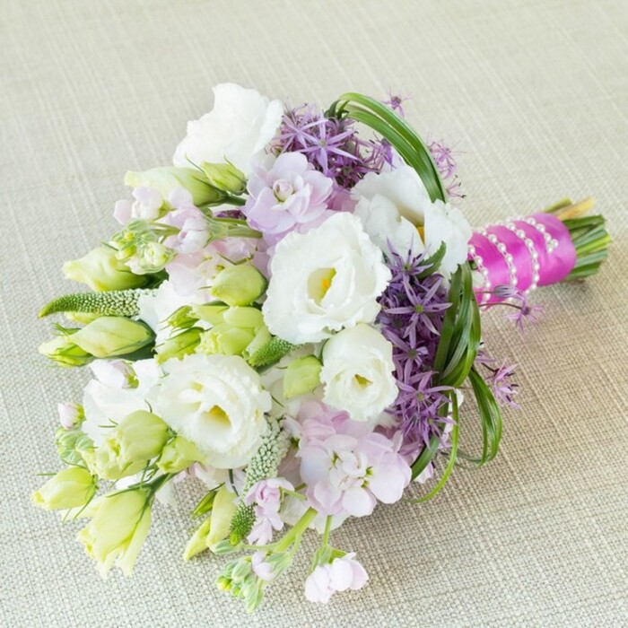 Placed diagonally across a soft cream linen reminiscent of Canonbury's artisanal style, this vibrant bouquet is a testament to refined local craftsmanship. The focal point comprises several grand white Lisianthus (Eustoma) blooms, their ruffled petals and pale yellow-green centers exuding freshness and purity-much like the spring blossoms in nearby Canonbury Gardens. These central white flowers are playfully complemented by clusters of lavender and pale pink stock, which add gentle notes of pastel harmony and fragrance that invite reminiscence of early morning strolls past hidden squares. Punctuating the arrangement, deep purple ornamental alliums present bold, star-shaped florets that introduce depth and structured contrast, much in the way Canonbury balances the historic and the contemporary. Slender green foliage, feathery and bright, weaves through the bouquet, with buds of varying sizes hinting at natural growth and vitality. All is bound together with a striking fuchsia-magenta ribbon, symbolising celebration, and adorned with an artful strand of lustrous pearls-details that echo the understated elegance Canonbury Flowers brings to local weddings and sophisticated events. Whether for a bridal bouquet, an anniversary, or another cherished occasion, this arrangement infuses romance, joy, and the unique spirit of Canonbury into every gathering.