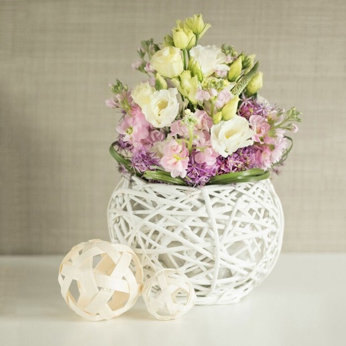 Elegance and warmth converge in this table arrangement, which captures the refined yet inviting spirit of Canonbury with every graceful detail. The composition features creamy white and gentle yellow lisianthus-petals hinting at the softness of morning light-standing proudly alongside tall, pastel pink matthiola that add a vertical, architectural touch, reminiscent of the area's grand Georgian homes. Purple filler blooms, perhaps miniature asters or alliums, nestle at the base, accentuating the bouquet's lushness and echoing the colourful abundance of a spring walk through Highbury Fields. The vibrant ensemble is set within a distinctive spherical vase, an open-work weave of white branches or rattan whose intricate craftsmanship beautifully balances lightness and structural interest. To the left, two decorative spheres made of pale natural wood or rattan mirror the vase's textural complexity, enhancing the coordinated, handmade feel. The clean, bright white tabletop and warm beige background, featuring subtle vertical fabric texture, create a sophisticated stage for the floral artistry, illuminated by gentle, diffused daylight. Ideal for stylish Canonbury weddings, refined brunches, or as a centrepiece in elegant townhouses, this image encapsulates the attention to detail, fresh local flowers, and harmonious design Canonbury Flowers is celebrated for.