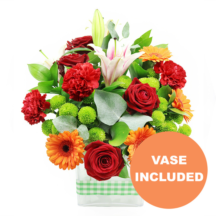 This image shows a joyful, colour-rich bouquet arranged in a contemporary clear glass vase, captured against a stark white background that lets every petal stand out. The composition is generously full and rounded, dominated by warm, festive tones of red and orange with lively splashes of bright green and serene white. Several deep crimson roses, their velvety petals tightly layered in classic spirals, anchor the design and bring a sense of timeless romance. Nestled alongside are red-orange carnations, whose frilled edges form soft, rippling textures that contrast beautifully with the smoother rose petals. Scattered throughout the bouquet, radiant orange gerbera daisies with dark, almost black central disks radiate like miniature suns, their broad, flat petals contributing a bold, graphic element that suits modern Canonbury apartments and studios. Rising through the centre and slightly off to one side, tall white Oriental lilies provide graceful height; some are fully open with delicate pink veins glowing along the length of each trumpet-shaped petal, while others remain in tight, creamy buds, hinting at freshness and longevity. Between these larger blooms, clusters of small, spherical lime-green pompom chrysanthemums add playful pops of colour and a pleasantly bouncy texture, softening the transitions between flowers. Rich greenery frames everything: broad, glossy green leaves and sprays of distinctive silver-green eucalyptus weave through the arrangement, creating depth and a gentle sense of movement, like a breeze running through a garden along Canonbury Park. The stems are gathered into a sleek, rectangular glass vase, its straight lines contrasting the organic forms above, and a light green and white gingham ribbon is neatly wrapped around its base, adding a homely, cheerful detail. In the lower right corner of the photograph, an orange circular badge with bold white capitals declares VASE INCLUDED, overlapping the vase just enough to signal that this polished, ready-to-display container is part of the complete floral gift.
