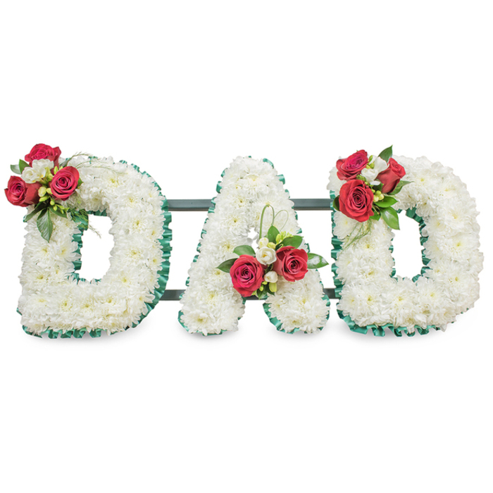 A poised and solemn floral tribute spells DAD in three substantial block letters, each letter meticulously covered in tightly packed, pristine white chrysanthemums that give a uniformly soft, pillowed texture across the surface. The outline of every letter is finished with a deep emerald, ribbed ribbon that provides a crisp edge and structural clarity against the white field. Artful clusters punctuate the composition: the first D bears a prominent spray of deep red roses mingled with small white freesia and glossy green foliage on its upper curve; the A displays a complementary arrangement positioned within its left diagonal near the base, accompanied by a delicate sweep of bright green foliage; and the final D mirrors the first with another matching bouquet of red roses, white freesia and abundant leaves. Subtle dark metal support bars run horizontally behind the letters, visible only as shadowed lines that stabilise the installation for transport and display at a funeral or memorial service. Under even, soft lighting the chrysanthemum petals read as gentle clouds while the roses lend velvety, tender accents - a combination intended to convey devoted love and lasting respect. I envisage this piece being chosen for a service near Canonbury's small chapels or placed beside a graveside plot close to Islington's quieter corners, where the colours and textures can speak plainly of grief, affection and remembrance.