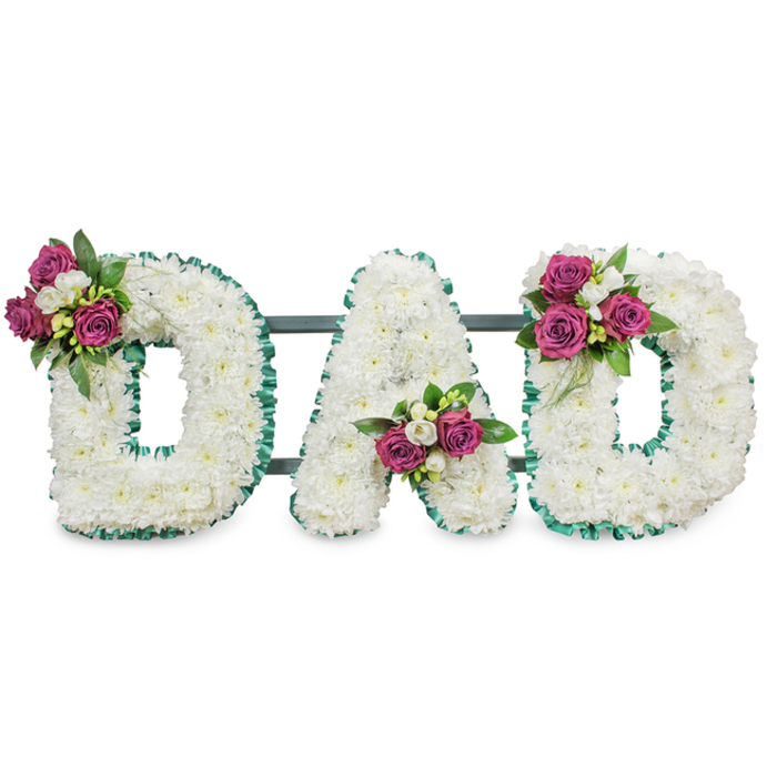 A lovingly crafted memorial arrangement spells the word DAD in large, sculpted capital letters, each letter formed from a full, cushiony mass of immaculate white chrysanthemums (or similar fluffy white blossoms) that create a seamless, soft surface. The letters are outlined with a vibrant emerald green ruffled ribbon that follows both the outer and inner edges, giving each character definition and a respectful, formal edge beneath gentle daylight-as you might see in a chapel near New River Walk. A slim, dark green or grey horizontal bar runs behind the three letters, discreetly linking them and providing a sturdy framework for display at a funeral or graveside. Small, precisely placed clusters of deep magenta-pink roses punctuate the composition: one cluster rests on the upper left curve of the first D, another nestles into the lower central V of the A, and a final matching cluster graces the upper right curve of the second D. Interspersed among the roses are tiny white freesia or similar delicate fillers, offering a faint, sweet fragrance and a fine contrast to the chrysanthemum heads, while lush dark foliage anchors the palette. As Canonbury's own florists would arrange, this tribute balances quiet dignity with personal warmth-crafted for those who wish to honour a father with a clear, heartfelt statement of remembrance and love at a local service or memorial.
