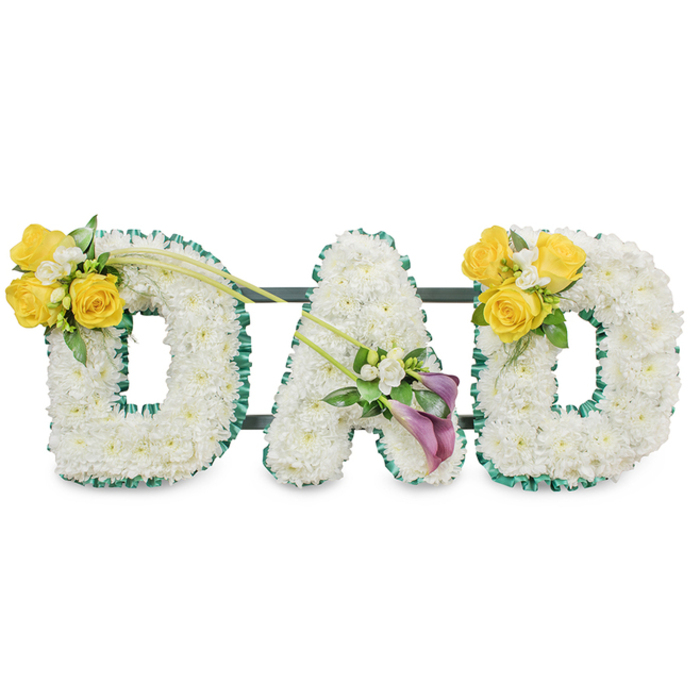 A striking, handcrafted memorial spelling DAD in three horizontal, freestanding letters, each letter formed from a dense bed of small white chrysanthemums that give the shapes a soft, rounded fullness. The perimeter of each letter is neatly trimmed with a ruffled dark green satin ribbon or tightly clipped foliage, the rich border creating a crisp visual edge against the white blooms and a dignified, almost tailored finish. Behind the trio, slim dark-green metal bars are visible, connecting and supporting the letters so they present as a single, cohesive tribute suitable for placement at a service or graveside. The left D is adorned on its upper left curve with a bright cluster of yellow roses - about three to four blooms - nestled among small white freesia accents and glossy green leaves; a couple of long, arching green stems gently extend from this cluster to add movement across to the central letter. The A carries a smaller, refined grouping on its lower diagonal composed of two deep purple calla lilies surrounded by delicate white fillers and fresh foliage, their sculpted forms offering a moment of quiet devotion. The right D mirrors the left with another sunny cluster of yellow roses and white freesia, balancing the composition. Symbolically, the white chrysanthemums speak of peace and purity, the yellow roses of affection and remembrance, and the purple callas of reverence. As Canonbury Flowers often arranges for families across Canonbury Square, St Pauls Road and nearby Barnsbury, this piece is presented with careful scent control, durable construction and a gentle, consoling visual language for saying goodbye.