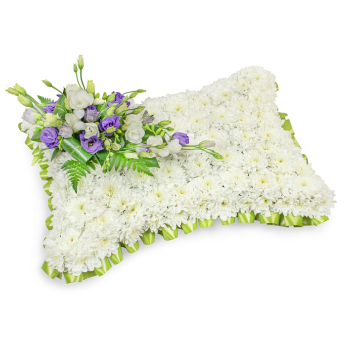 A tasteful rectangular sympathy pillow formed almost entirely of tiny, snow-white chrysanthemum heads, the flowers set so closely that the surface appears like a smooth, comforting pad of blossoms. The pillow's soft silhouette is outlined by a light green, pleated ribbon that follows the rounded corners in a careful scalloped trim, offering a gentle visual lift against the wash of white. In the upper-left quadrant a small but elegant spray anchors the design: white lisianthus bring soft, layered petals while deep purple lisianthus-some newly opened, some still in bud-introduce a quiet note of contrast and reverence. Interlaced fern fronds and glossy foliage add green texture and natural rhythm, and a darker green wrap at the stem base secures the cluster with a professional finish. The image is shot on a pristine white background with soft shadows that emphasise both the pillow's dimensional puff and the artisan placement of each chrysanthemum head. The overall effect is serene and composed, intended for memorial services, church viewings or graveside farewells, and it reflects the sympathetic style often requested by families around Canonbury and the surrounding Islington neighbourhood, including Essex Road and Upper Street. The arrangement suggests the hush of a chapel, a faintly green fresh scent and the reassuring competence of a local florist handcrafting a tribute meant to soothe and remember.