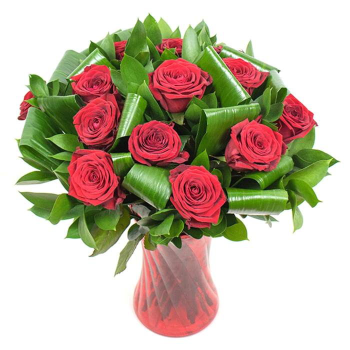 A sophisticated, deeply romantic bouquet composed of roughly a dozen richly hued crimson roses forms the visual heart of this image, each bloom open to a perfect, velvety cup of tightly spiralled petals. The roses are densely gathered to create a lavish, rounded silhouette, their passionate red intensified by even, soft lighting that sculpts every petal edge and shadow. Nestled among the rose heads is an abundance of dark green foliage: slender glossy leaves that build volume and larger, bright emerald leaves that have been expertly folded and rolled into cylindrical shapes and graceful spirals. These architectural green elements are purposefully placed to cradle the roses and introduce a modern, sculptural quality to a timeless composition. Everything is presented in a tall, translucent ruby-red glass vase with a subtle twisted rib texture; the vase's vertical lines catch and refract light, harmonising with the roses' deep tones while providing an elegant, stable base. Set against a stark white background, the arrangement becomes a study in contrast and craftsmanship - the polished greens, the velvety reds, the glass's faint undulations all visible in crisp detail. As arranged by Canonbury Flowers for clients across Canonbury, Essex Road and nearby Highbury Fields, this bouquet reads as a luxurious gift or centerpiece that communicates ardour and refined taste, ideal for anniversaries, Valentine's Day or any intimate celebration where fresh scent and expert floral design matter.