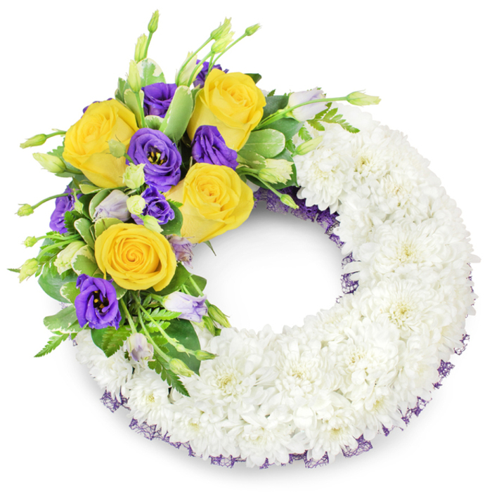 A meticulously arranged circular wreath, composed primarily of tightly packed white spray chrysanthemums that together form a plush, cloud-like band against a pristine white background. The chrysanthemums' many small, overlapping petals create a uniform texture and a sense of serene continuity around the open centre. On the upper left quadrant a graceful accent cluster arcs gently into view: honeyed yellow roses, fully opened with layered, buttery petals that radiate gentle warmth, paired with deep royal purple lisianthus (Eustoma) whose ruffled, velveteen blooms add a stately, contemplative contrast. Sprigs of pale lavender lisianthus buds and tender green shoots punctuate the focal spray, giving it natural rhythm, while varied foliage - glossy rounded leaves, faintly variegated greenery and fine fern fronds - provides structure and a soft, living backdrop. A subtle dark purple crinoline or mesh ribbon peeks from beneath the chrysanthemums at the inner and outer edges, a trim detail that quietly echoes the purple flowers and unifies the design. Bathed in soft, neutral light, the wreath reads as a dignified sympathy tribute, suited to services at local Canonbury chapels or for placing at a service in Barnsbury or near Highbury Fields. As described by a Canonbury-based florist at Canonbury Flowers, the arrangement balances purity and colour to offer a message of peace, remembrance and heartfelt farewell for bereaved families in the neighbourhood.