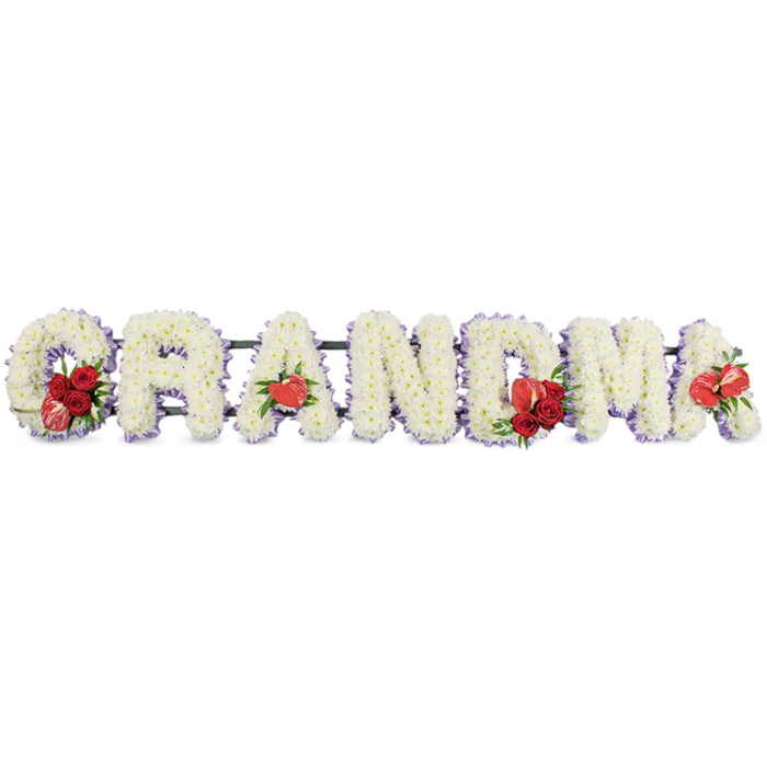 Set against a clean white backdrop, a lovingly crafted funeral flower arrangement spells out GRANDMA in bold, upright block capitals, each letter composed entirely of pristine white chrysanthemums or similar compact florets. The blossoms are packed so densely that they form a plush, velvety surface, catching the light with tiny glints on each petal tip. Around the perimeter of every letter runs a subtle border of lilac-tinged ribbon or backing, lending a gentle lavender glow and softening the otherwise stark white palette. The letters are aligned closely together in a straight horizontal line, all supported by a sturdy dark green metal or wooden stand that is only slightly visible from behind, ensuring that the word seems to float in front of the viewer. Deep red accents punctuate this sea of white at thoughtful intervals: on the first letter, G, a small arrangement of rich red roses, glossy foliage, and a variegated red-and-white calla-type flower nestles into the curve, drawing the eye to the beginning of the name. The first A is graced with a single red anthurium, its heart-shaped spathe echoing the love that the family feels. Toward the centre, the M carries another spray of red roses paired with a second anthurium, while the final A repeats this anthurium motif, completing the visual rhythm. The overall effect is one of calm reverence and personal devotion, the sort of tribute a Canonbury family might choose for a service at a nearby church or chapel, or for a graveside farewell in one of the local cemeteries, as a clear, floral statement of gratitude for a grandmother's life.