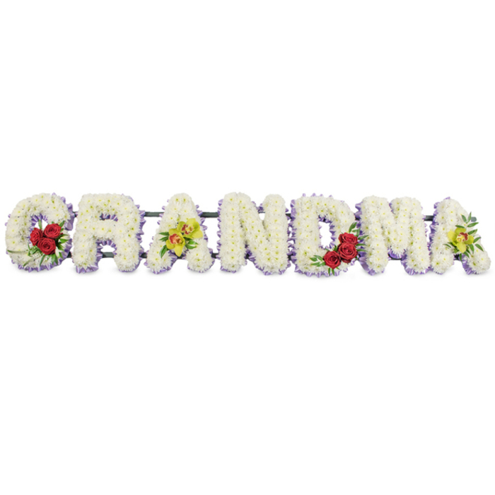 The photograph presents a horizontal floral word tribute spelling GRANDMA, meticulously arranged so that each capital letter appears full, three-dimensional, and gently sculpted from masses of fresh white blooms. These are most likely small-headed chrysanthemums, selected for their durability and consistent shape, and they are packed so tightly that the surface of each letter looks like a smooth field of snow, with individual petals only visible on closer inspection. Beneath this layer of white flowers, a subtle collar of light lavender blossoms, possibly statice or mini asters, edges the base of each letter and peeks out just enough to define the contours in a soft halo of purple. The florist then adds thoughtful touches of colour that personalise the tribute in a way many Canonbury families request when honouring a beloved grandmother. On the left-hand side, the G is embellished at its lower curve with three velvety red roses, tucked into a cushion of glossy dark foliage. Their deep crimson tones create a striking focal point, suggesting love and admiration. The first A features two unusual yellow-green orchids placed along the crossbar, their speckled centres and firm petals introducing a subtle contemporary feel. The central letters R, N, and D remain purely white within their lavender outlines, embodying simplicity and calm. Moving towards the right, the M carries a second trio of deep red roses in its lower right section, echoing the earlier accent and balancing the design. The final A mirrors the first, with a matching duo of green orchids resting near its centre. All seven letters are mounted on a discreet dark metal frame that runs behind them, giving the word structure without drawing attention from the flowers. The clean white background and even lighting ensure that every petal and colour stands out clearly, evoking the feeling of a peaceful service in one of Canonbury's quiet chapels, where this tribute would serve as a central expression of remembrance and affection.